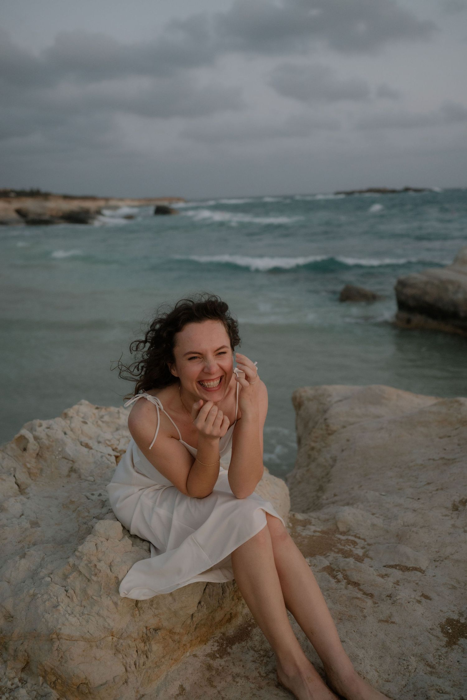 A smiling woman in a white dress sits on coastal rocks by a turquoise sea under a cloudy sky.