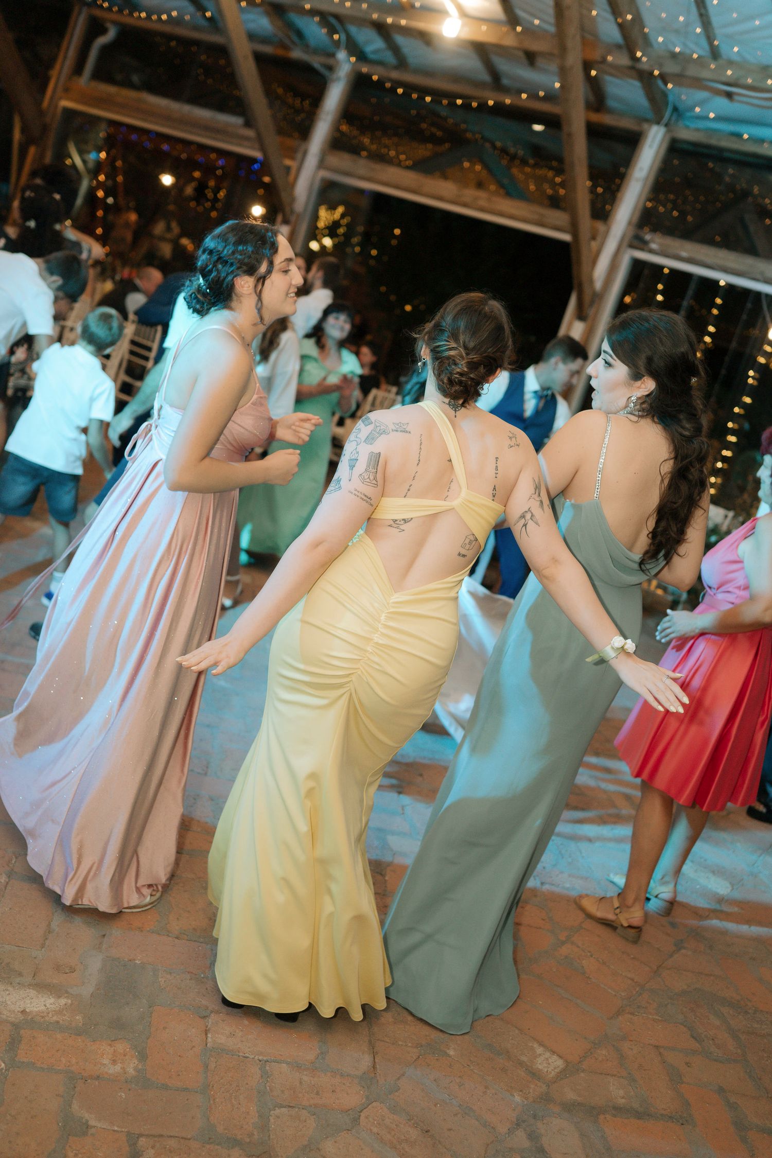 Wedding guests in pastel bridesmaid dresses dance together on a brick patio under twinkling string lights.