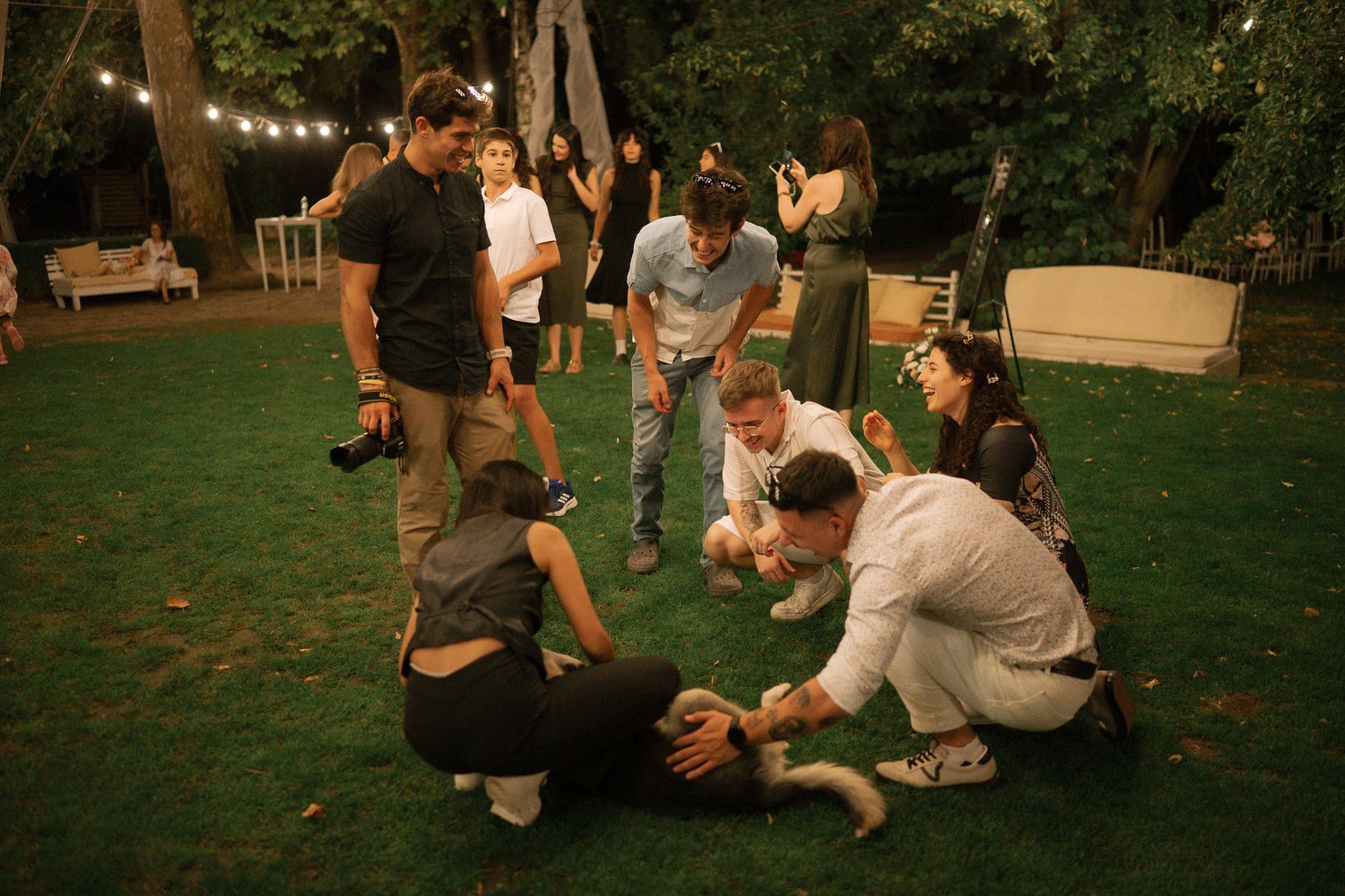 People gather on a green lawn at dusk playing a casual game while string lights glow in the background.