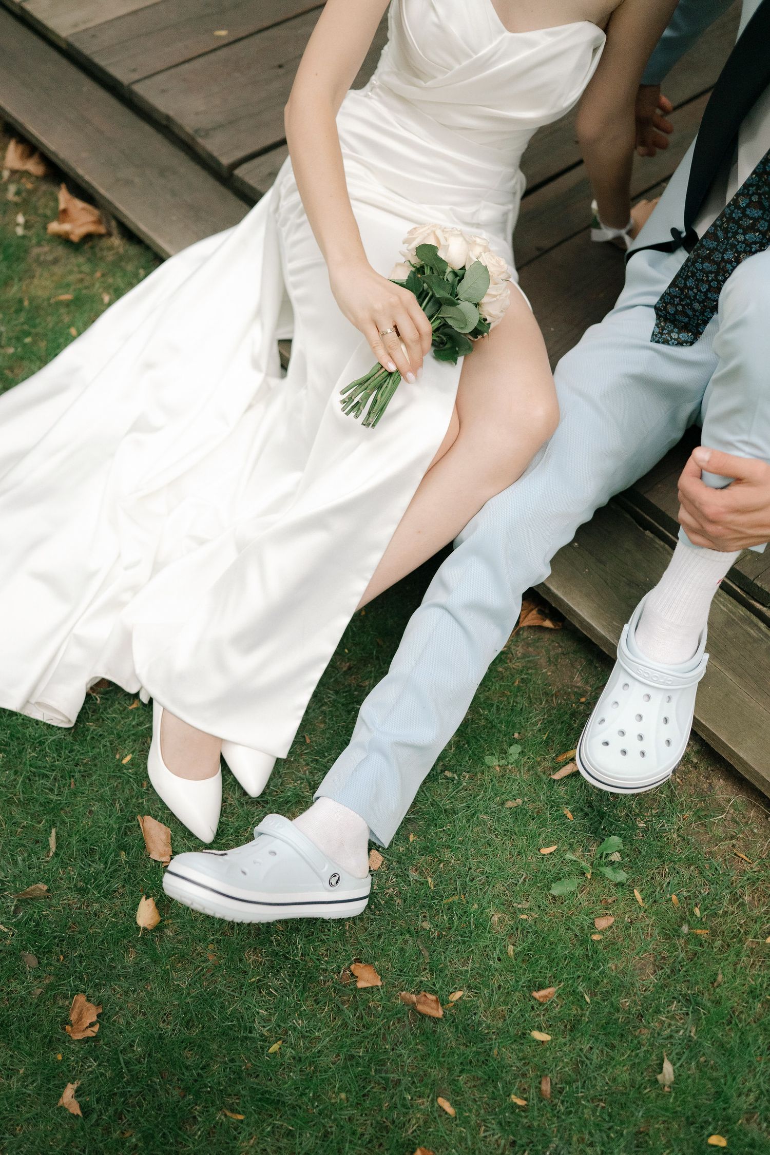 Close-up view of white crocs and wedding dress details on grass with scattered autumn leaves.