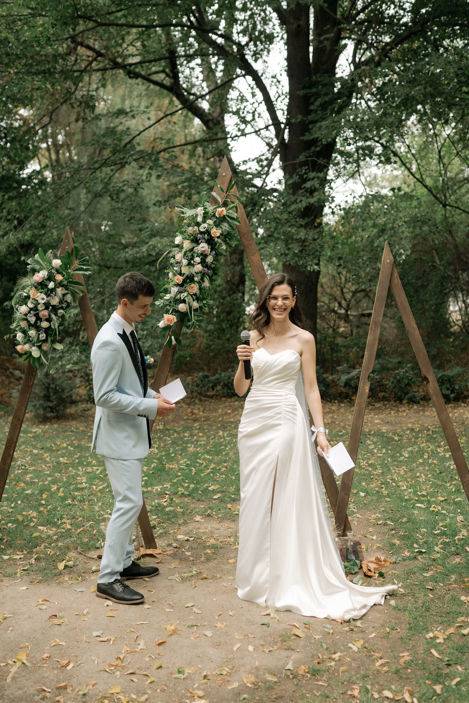A couple reads their vows under a wooden arch decorated with white floral arrangements in an outdoor garden setting.
