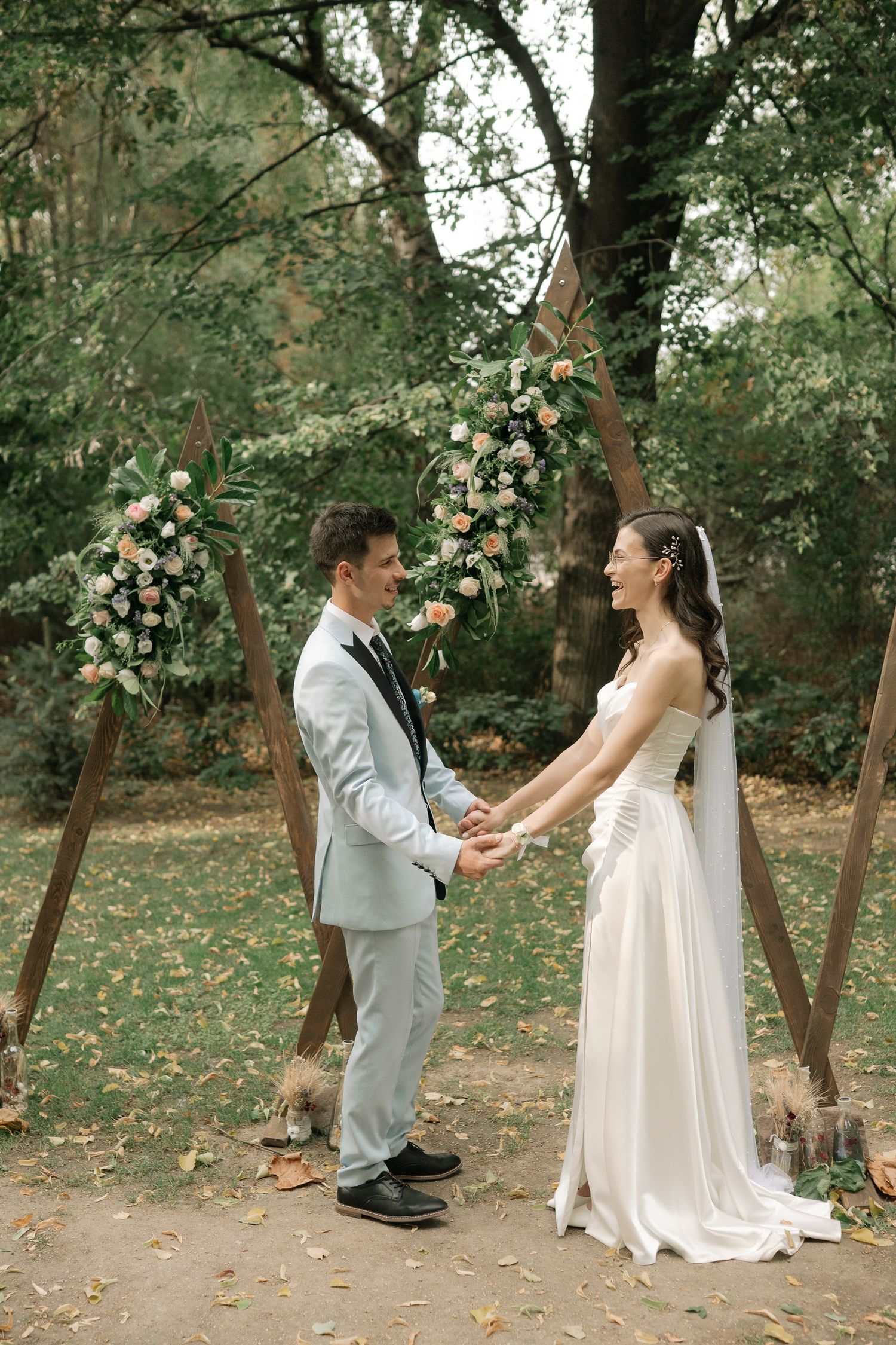 A couple in wedding attire dances playfully in a wooded outdoor setting with floral decorations visible in the background.