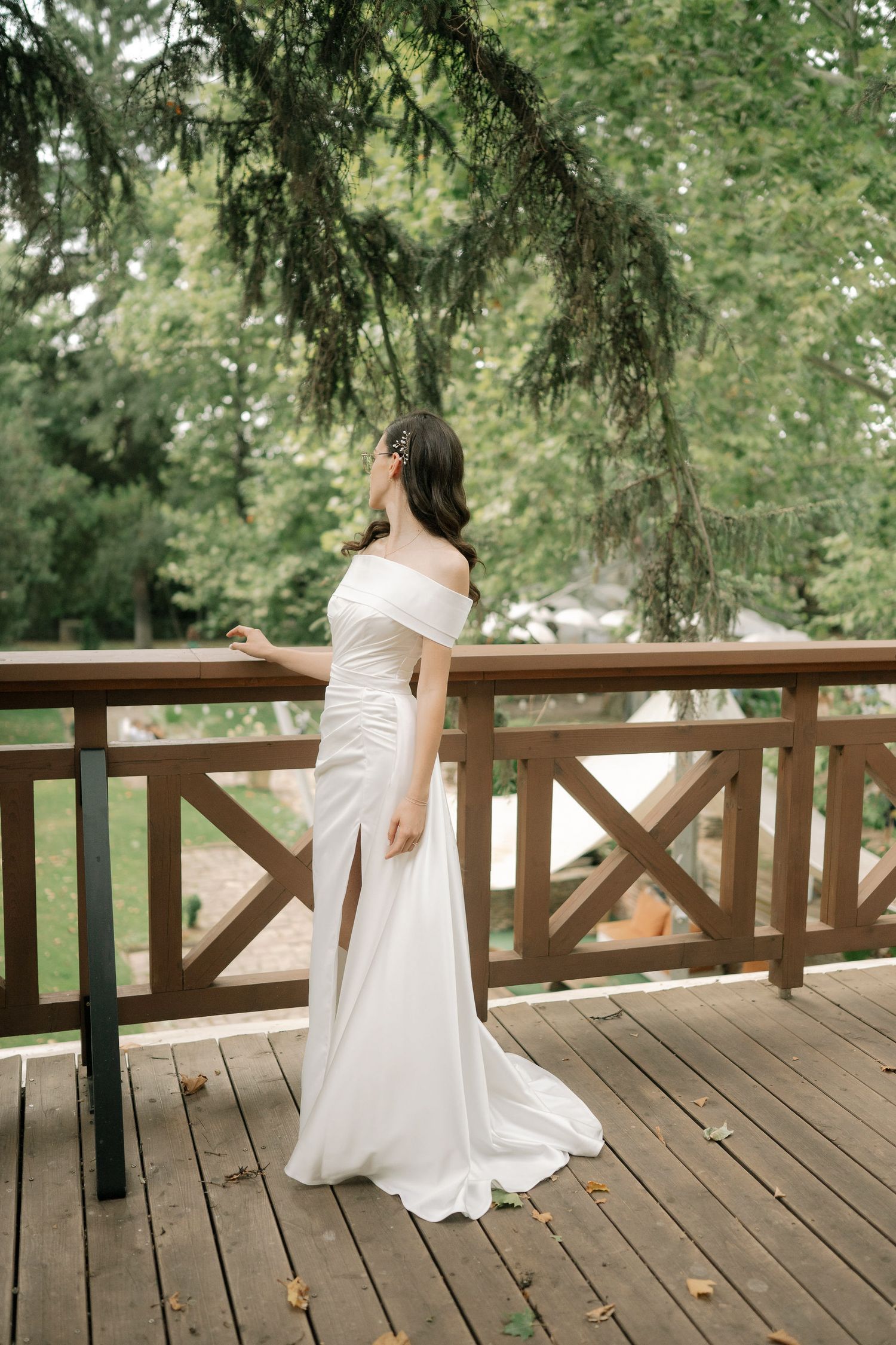 Elegant white off-shoulder wedding dress with side slit shown on wooden deck under overhanging evergreen branches.
