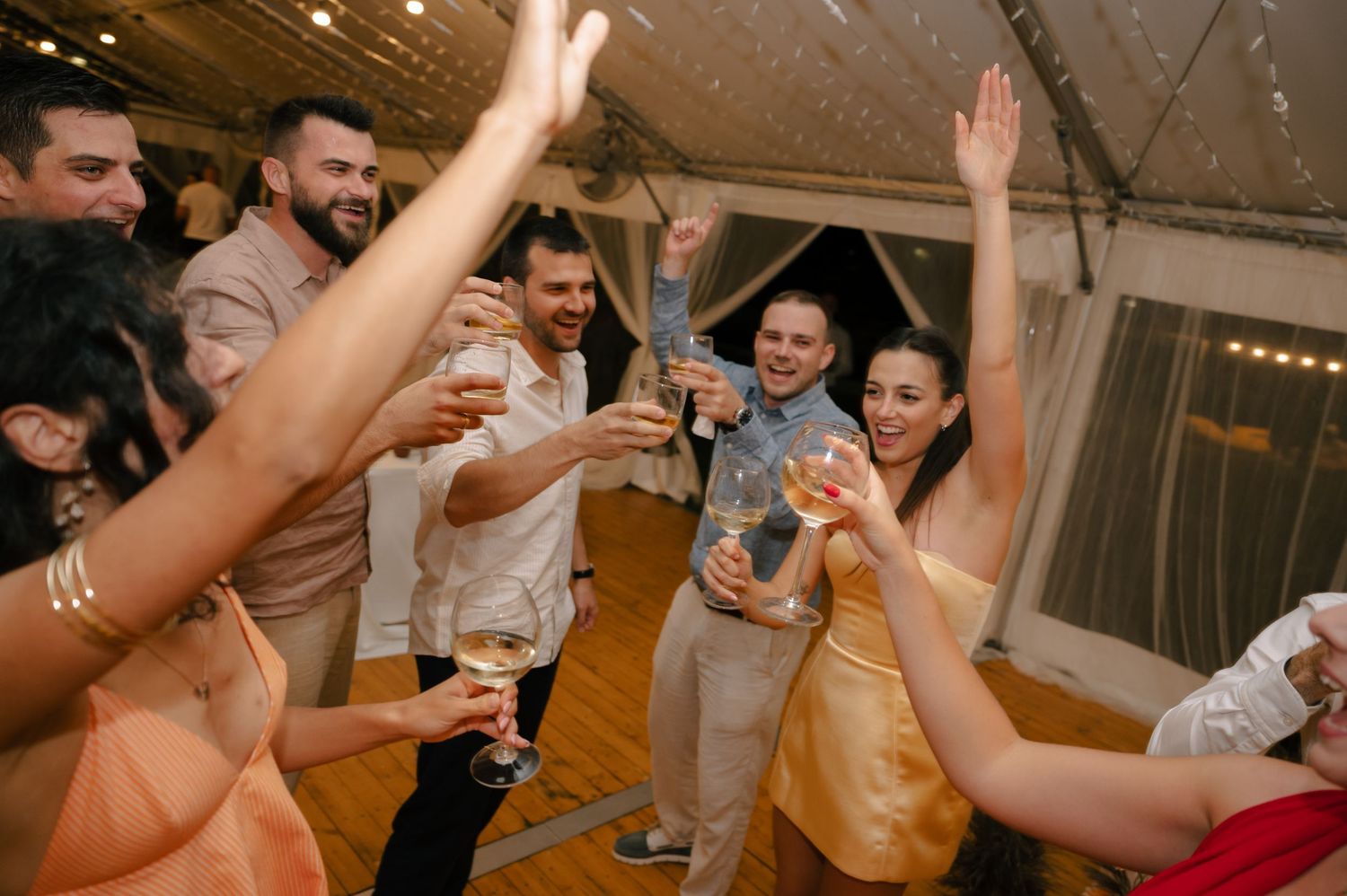 People dancing and celebrating at a wedding reception with raised hands and drinks on a wooden dance floor.