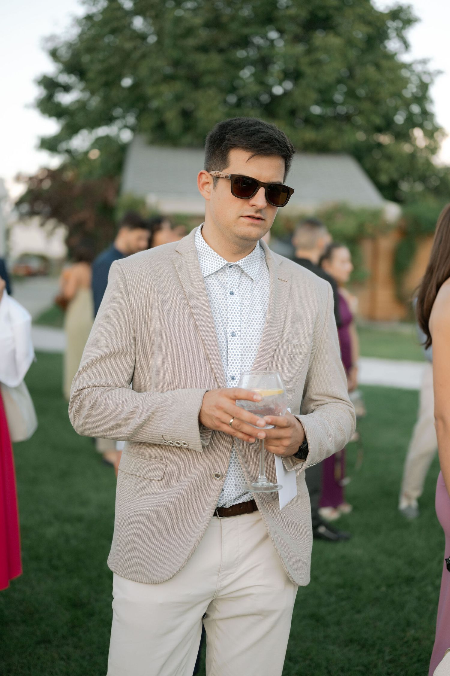 Groomsmen in beige suits and sunglasses check their phones while standing on grass during outdoor wedding event.