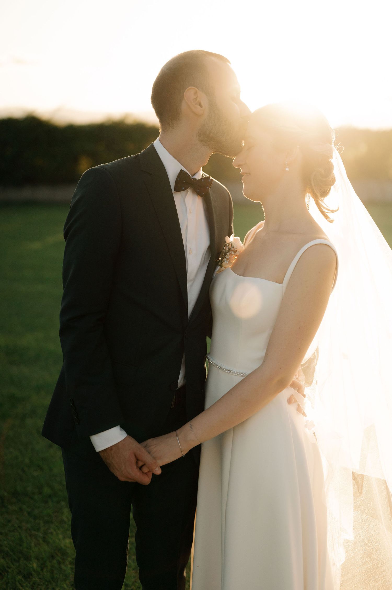 A romantic sunset wedding photo of a couple in formal attire holding hands in a grassy field with golden light behind them.