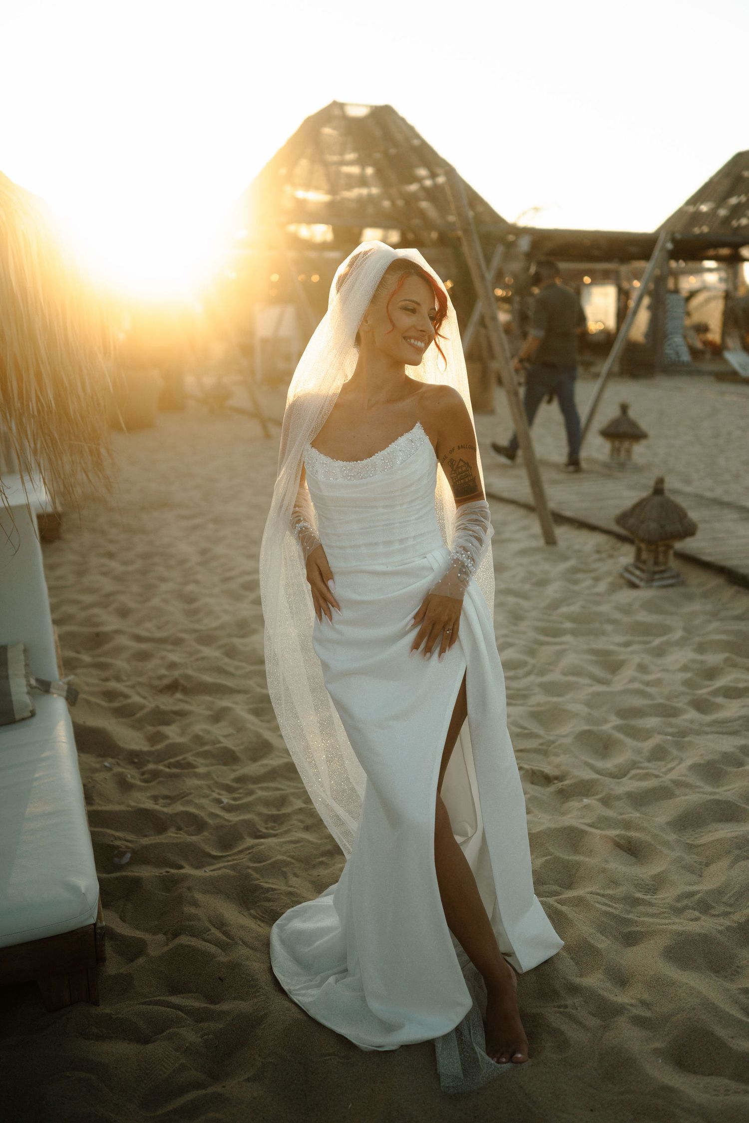 A person in a flowing white dress poses gracefully on a sandy beach at sunset.