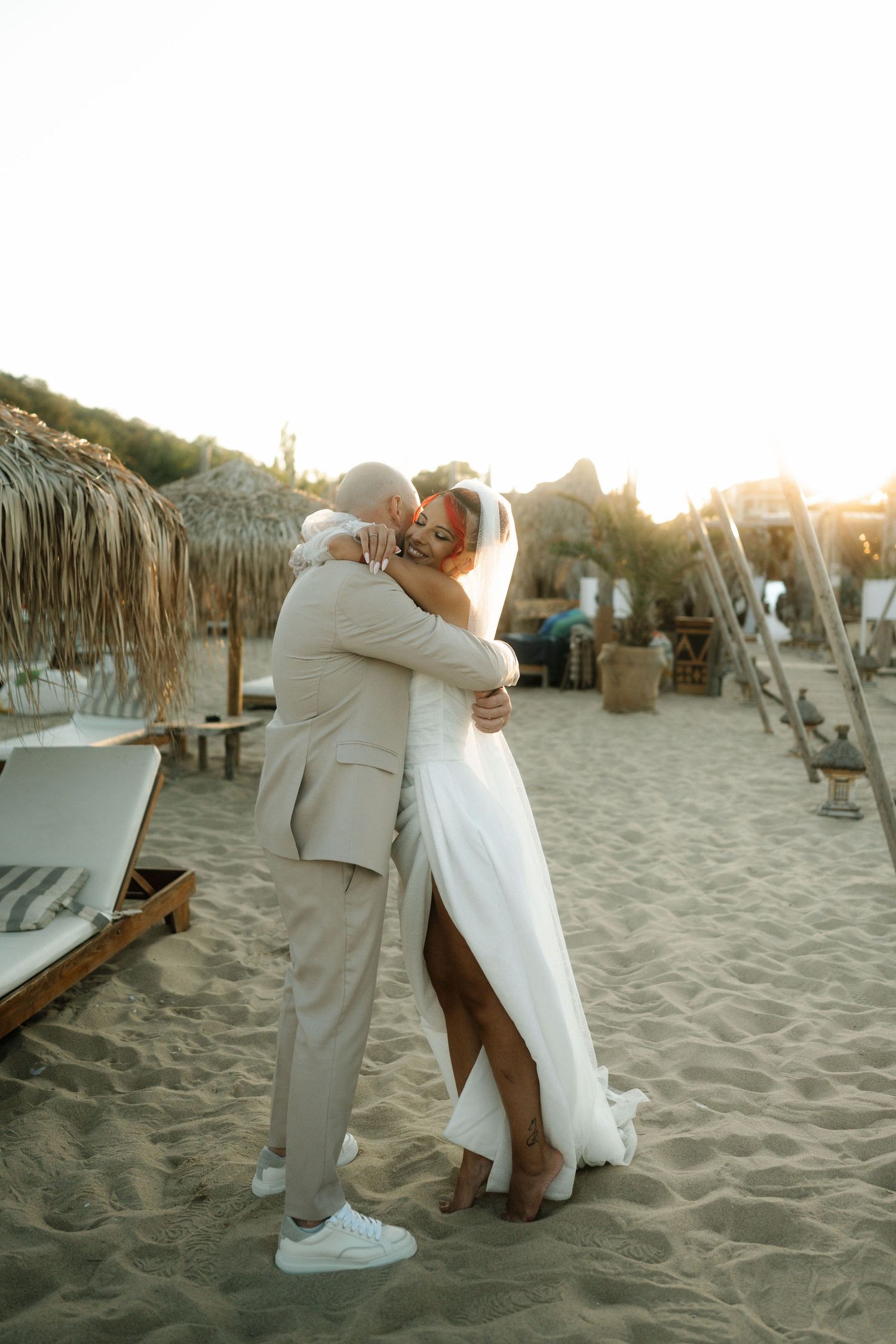 A couple embraces on a sandy beach at sunset with thatched umbrellas and beach decor in the background.