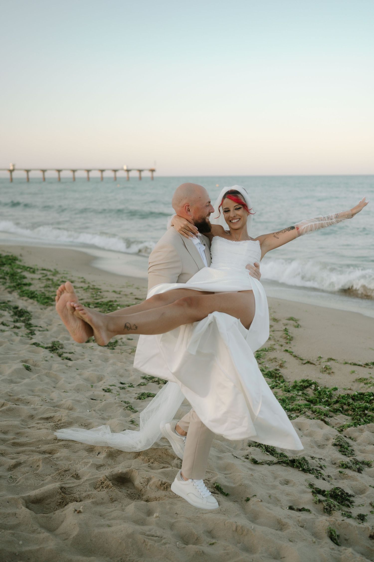 A couple in white attire dances joyfully on a sandy beach at sunset with ocean waves in the background.