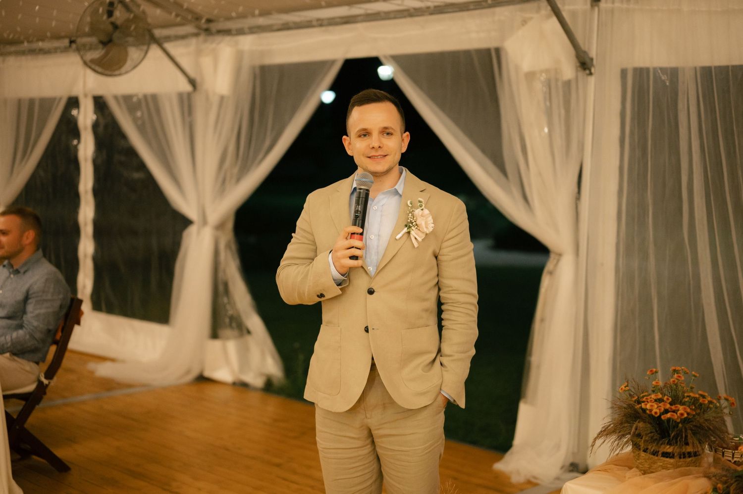 A speaker in a tan suit addresses wedding guests during an evening reception.