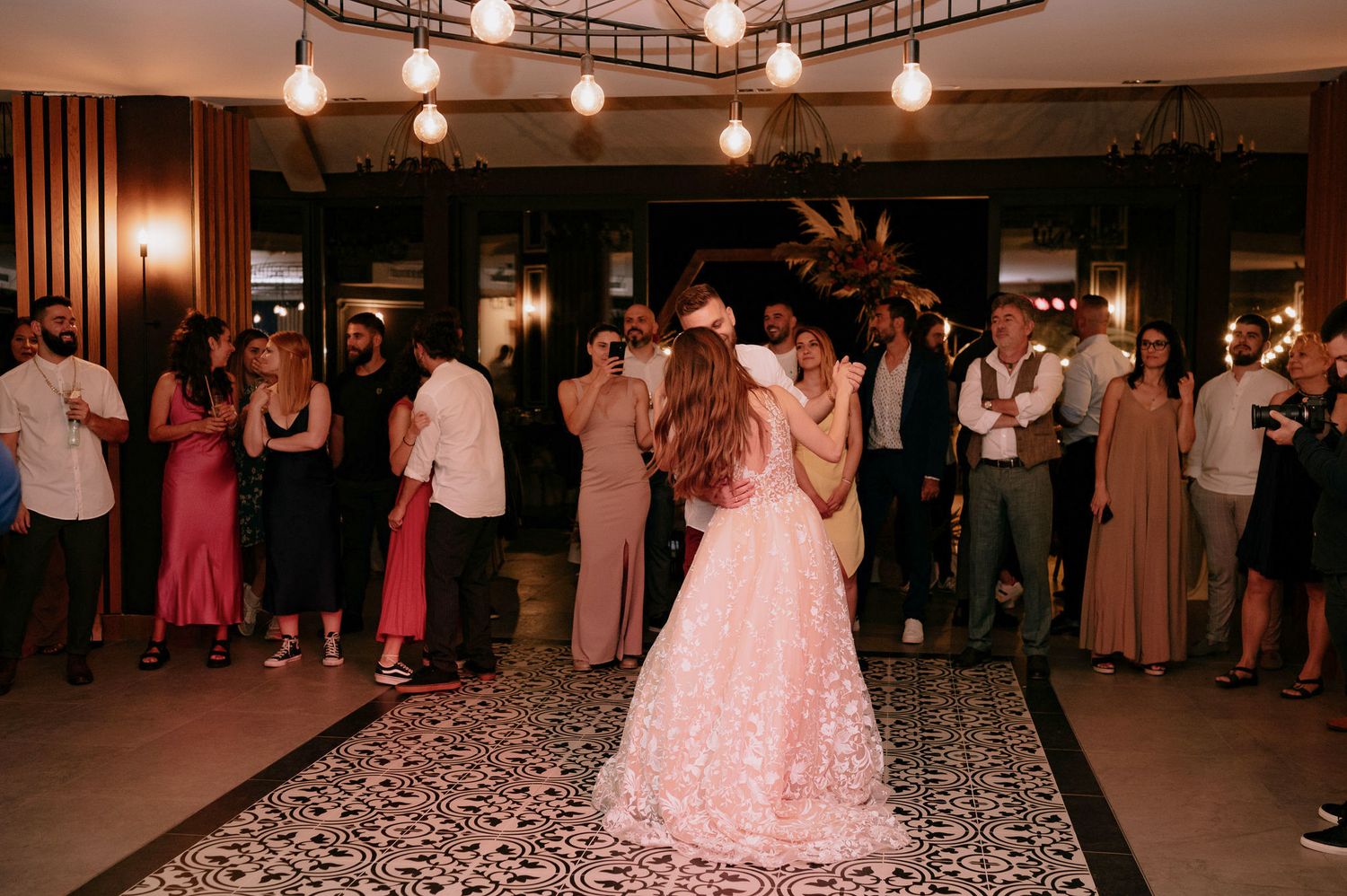 Wedding guests dance together in a dimly lit reception hall with string lights overhead and wood paneling on the walls.