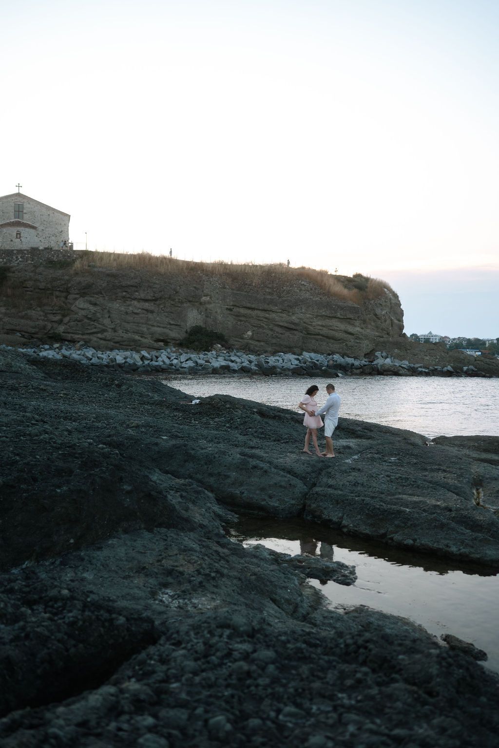 Two people stand on dark volcanic rocks near the ocean at sunset with a lighthouse visible in the distance.