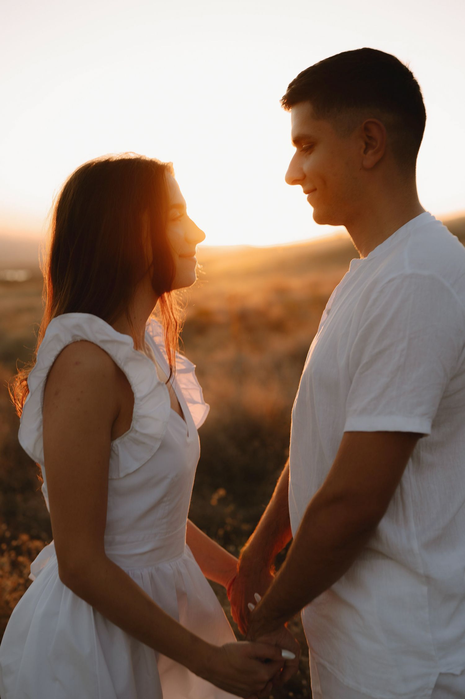 A romantic couple in white outfits embraces during golden hour sunset in a desert landscape.