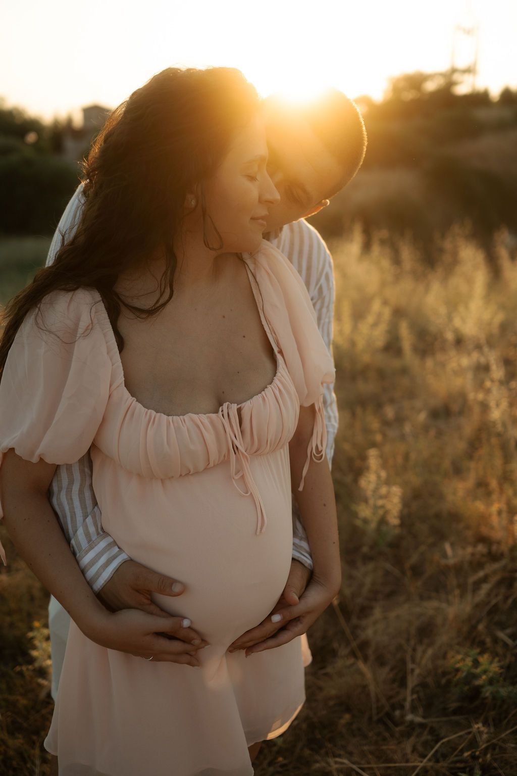 Maternity photos captured in a sun-drenched field with soft natural lighting and delicate pink dress.