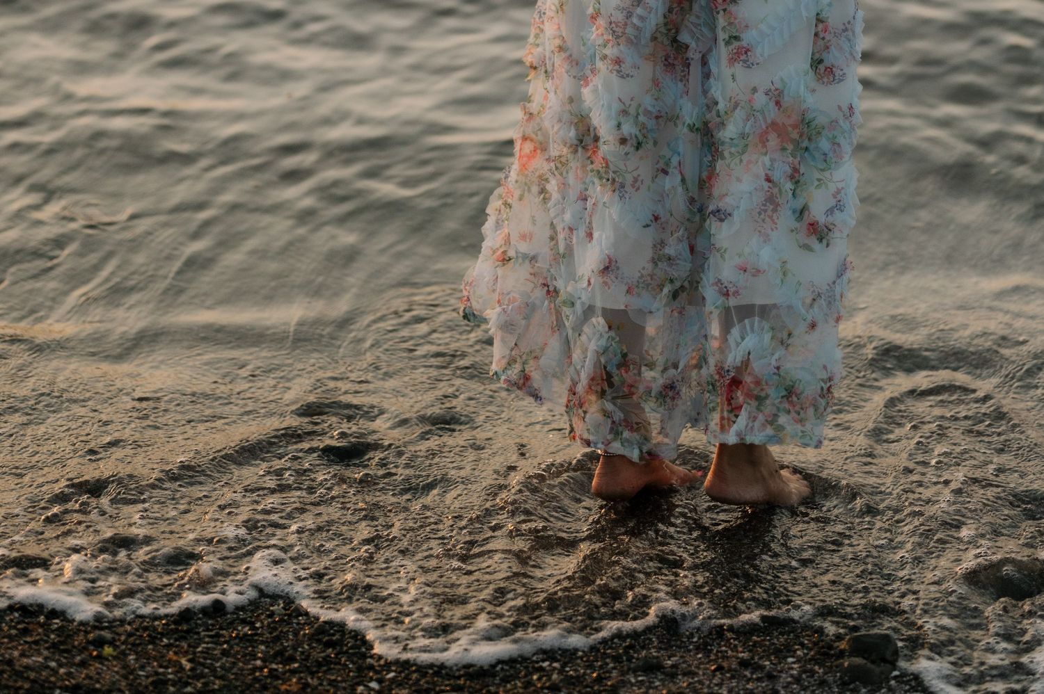 A person stands barefoot at the edge of a sandy beach as gentle waves lap at their feet in a floral dress.