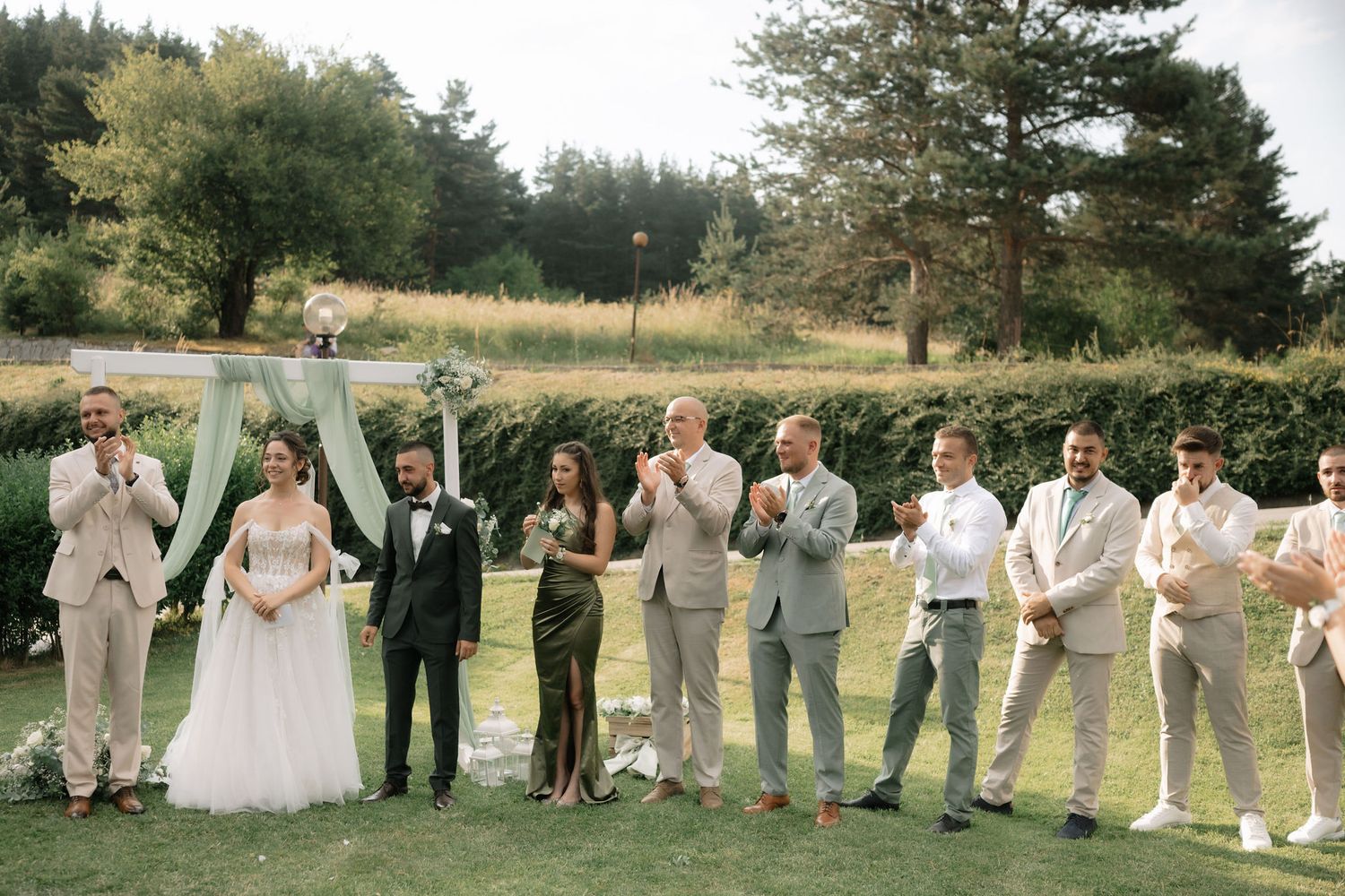Wedding party poses together outdoors in a line formation against a scenic natural backdrop of trees and grass.