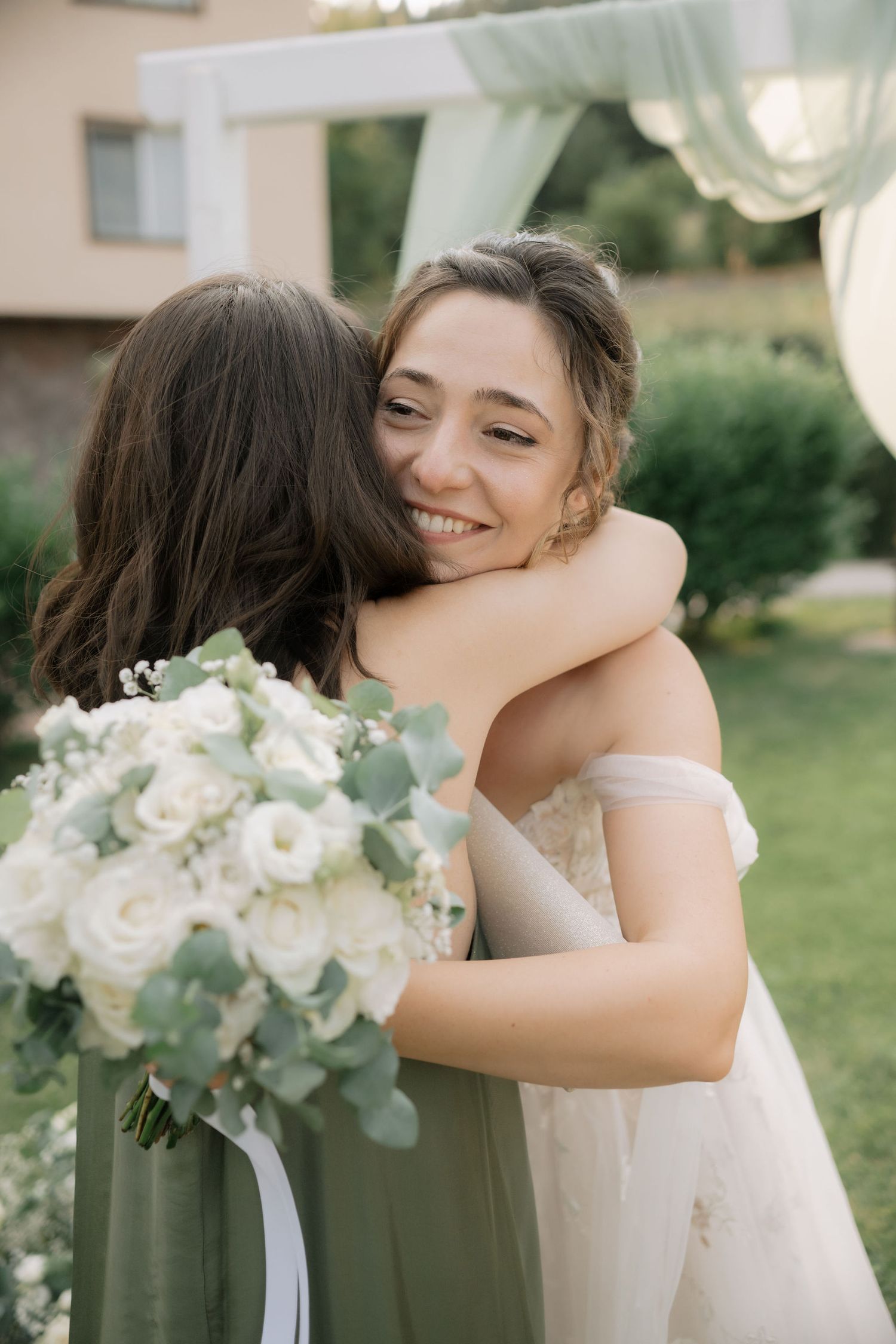 Two people embrace at a wedding celebration, with a beautiful white rose and eucalyptus bouquet visible.