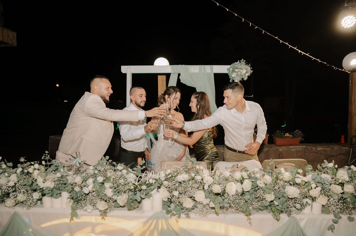 Wedding party enjoys a candlelit outdoor dinner reception under string lights with white floral decorations.