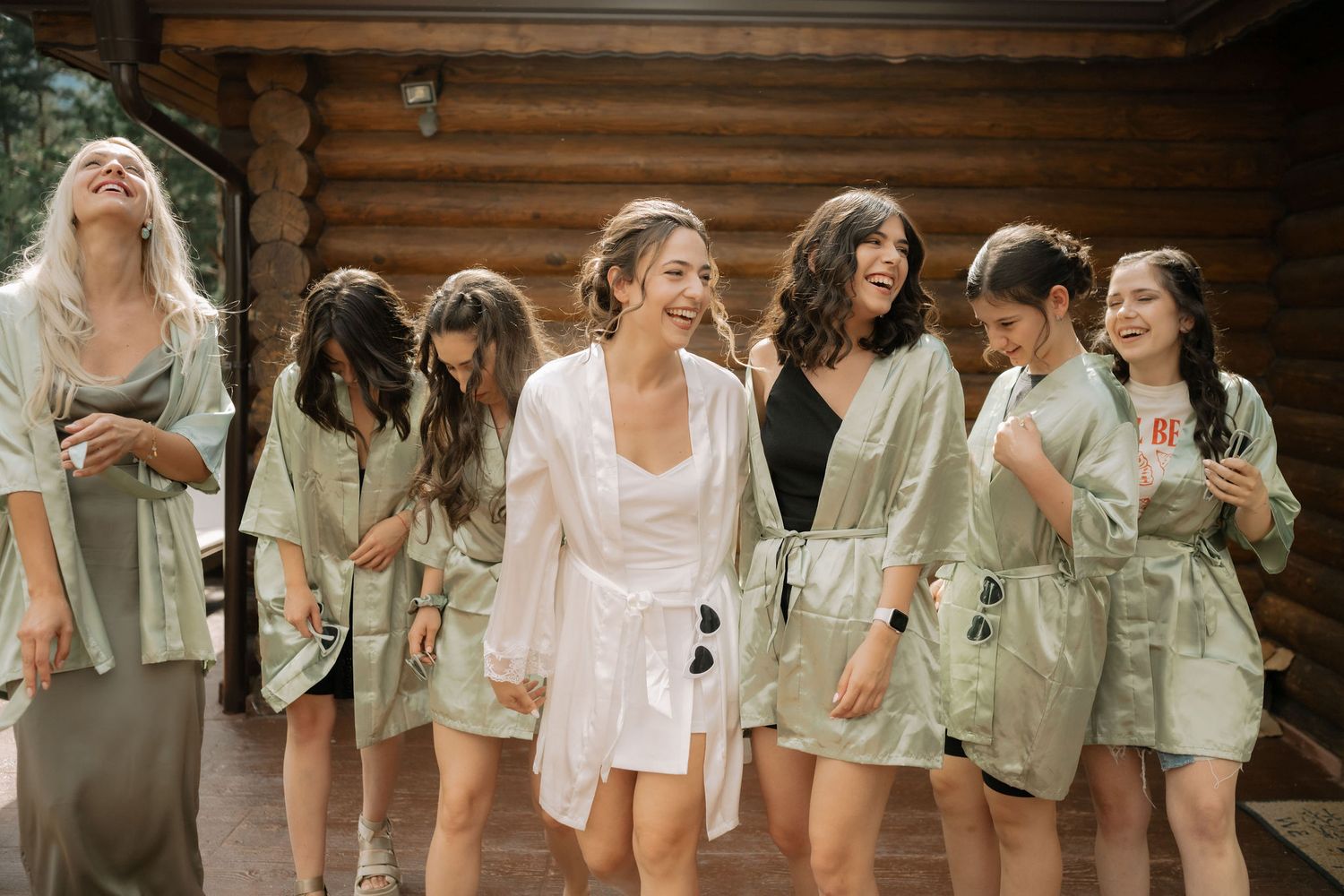Wedding party members in sage green dresses and white outfits gather together outside a rustic wooden cabin.