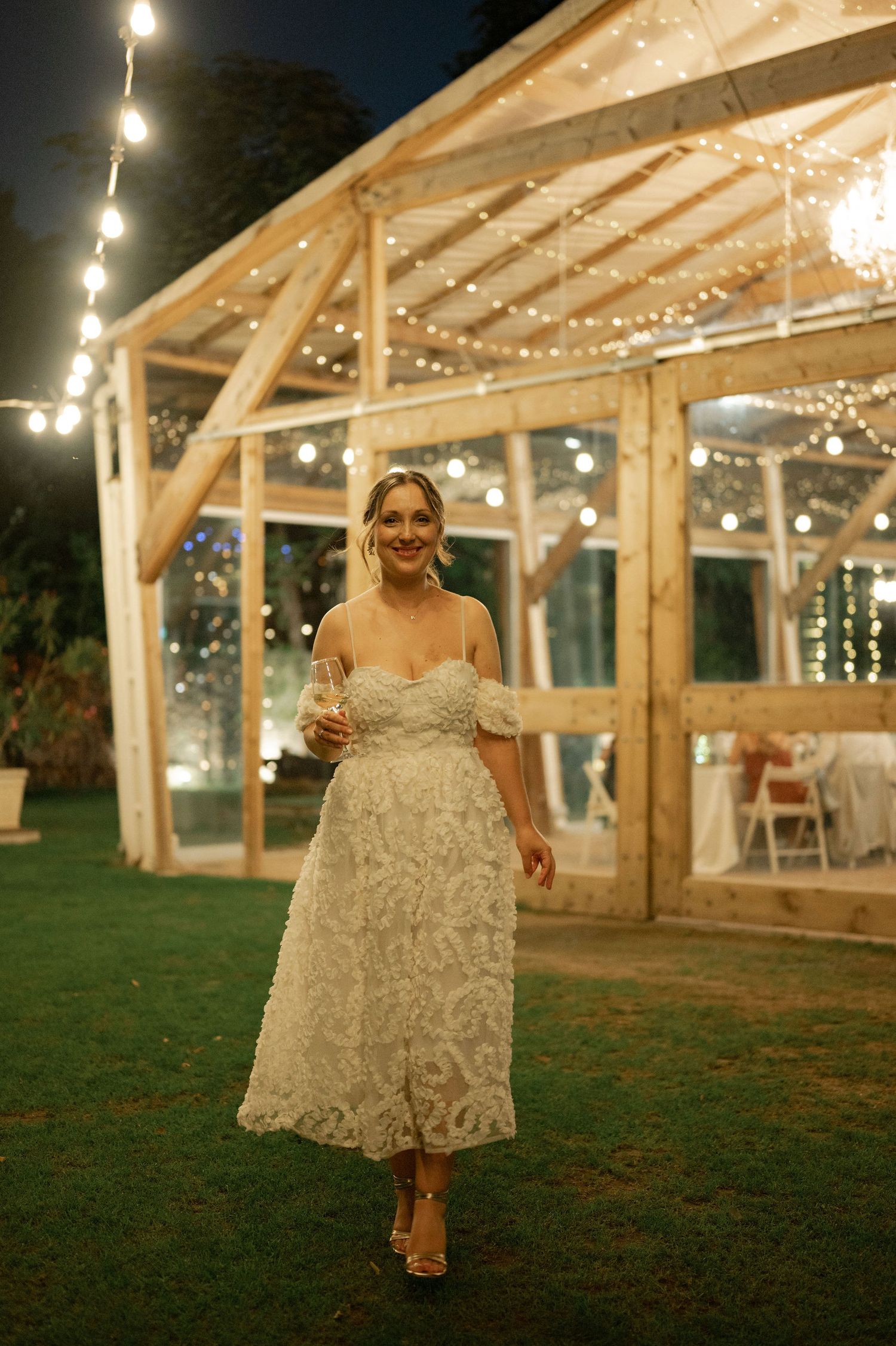 A beautiful wedding reception setup with string lights illuminating a rustic wooden barn structure and green lawn at dusk.
