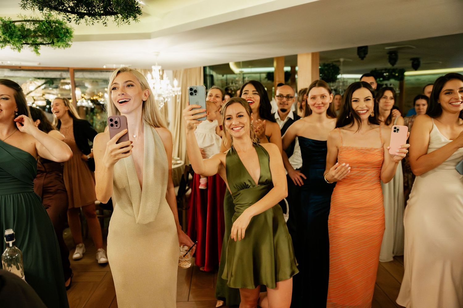 Wedding guests in colorful dresses dance and celebrate together at an indoor reception venue.