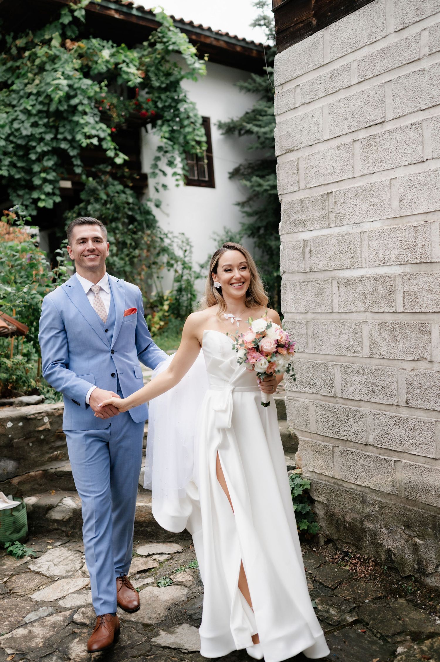 A couple in wedding attire walk up stone steps surrounded by greenery at a rustic venue.