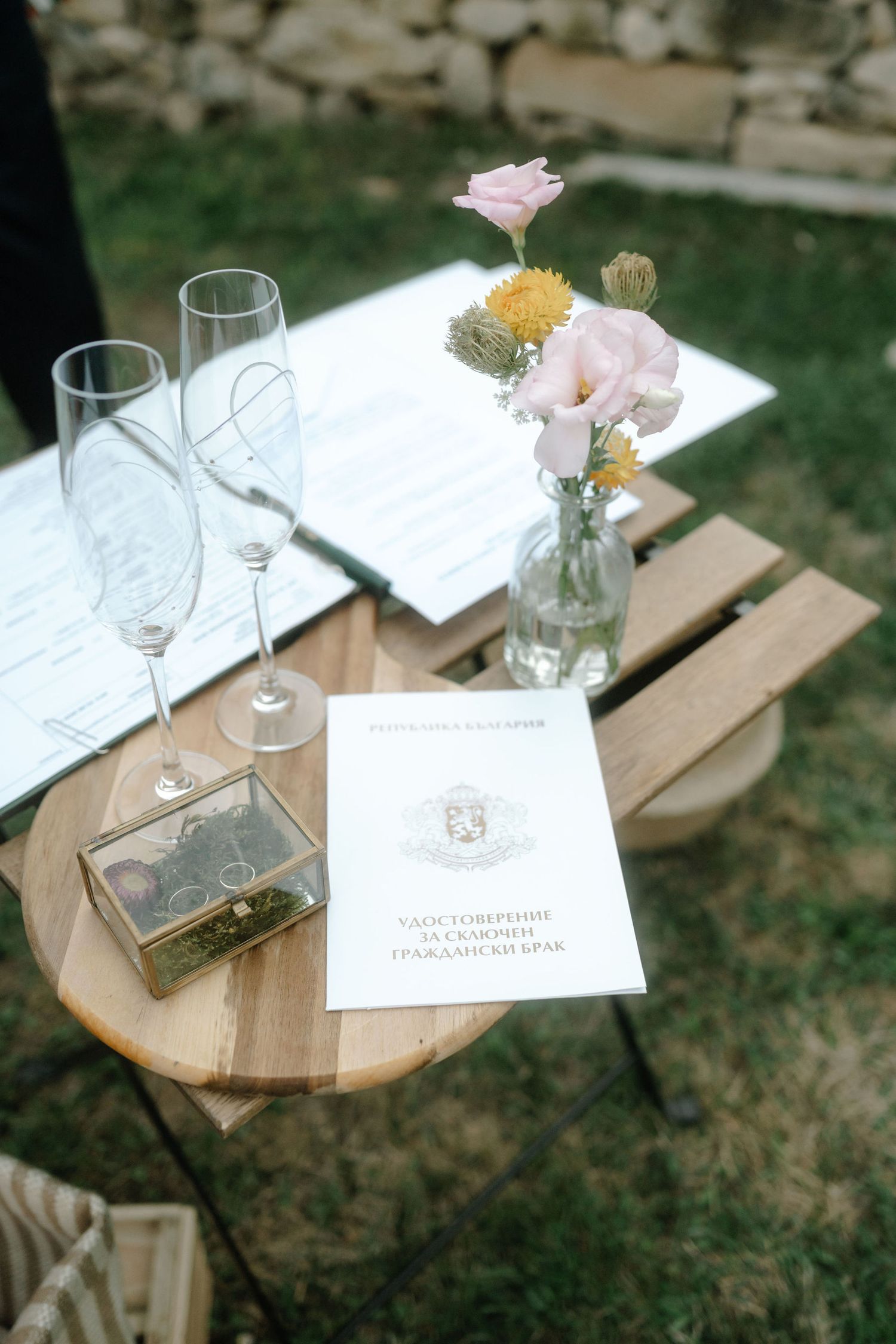 Wedding details with champagne glasses, delicate pink flowers in a vase, and a paper invitation on a wooden surface outdoors.