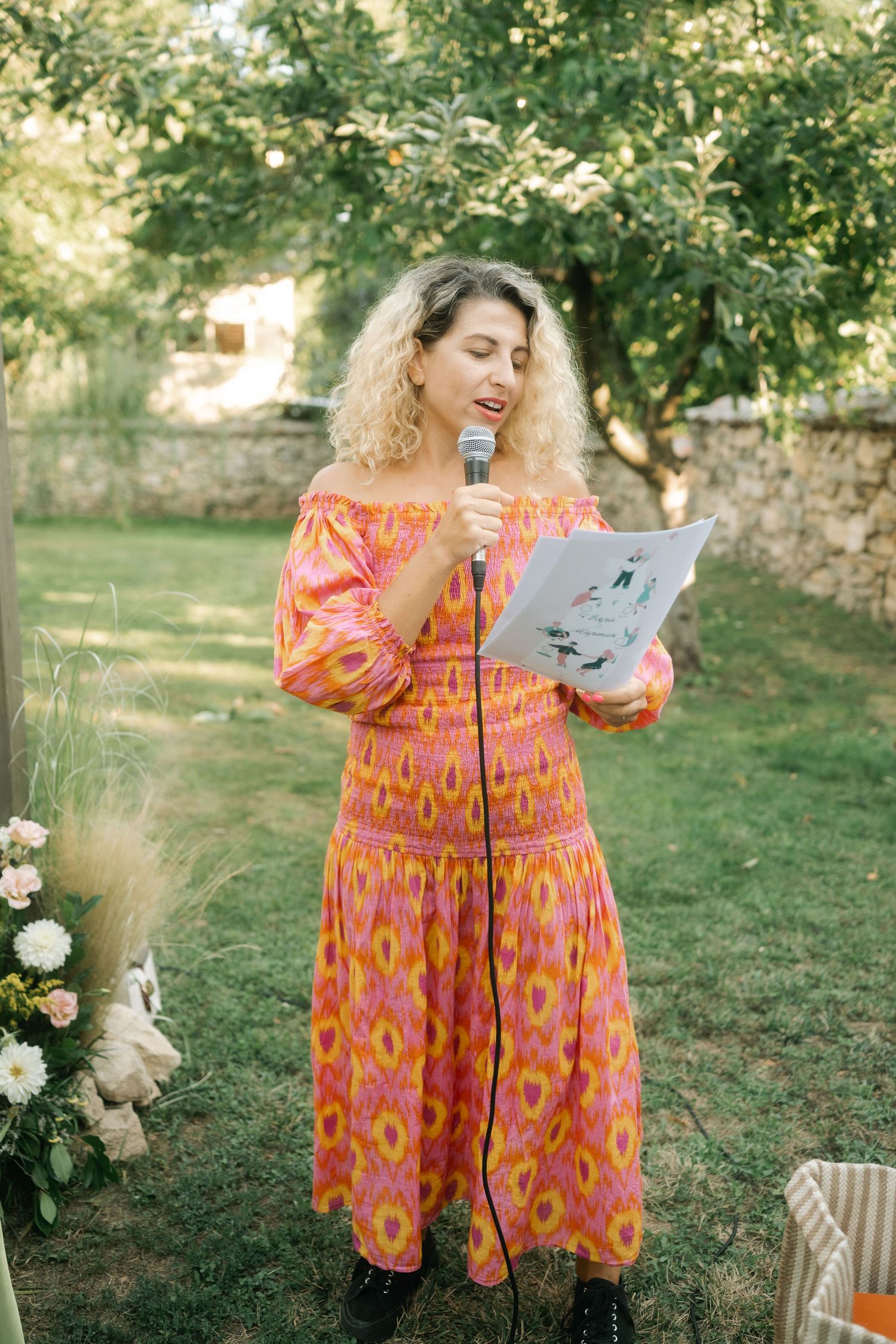 A person in a coral floral print dress reads from papers while standing in a garden setting with trees and flowers.