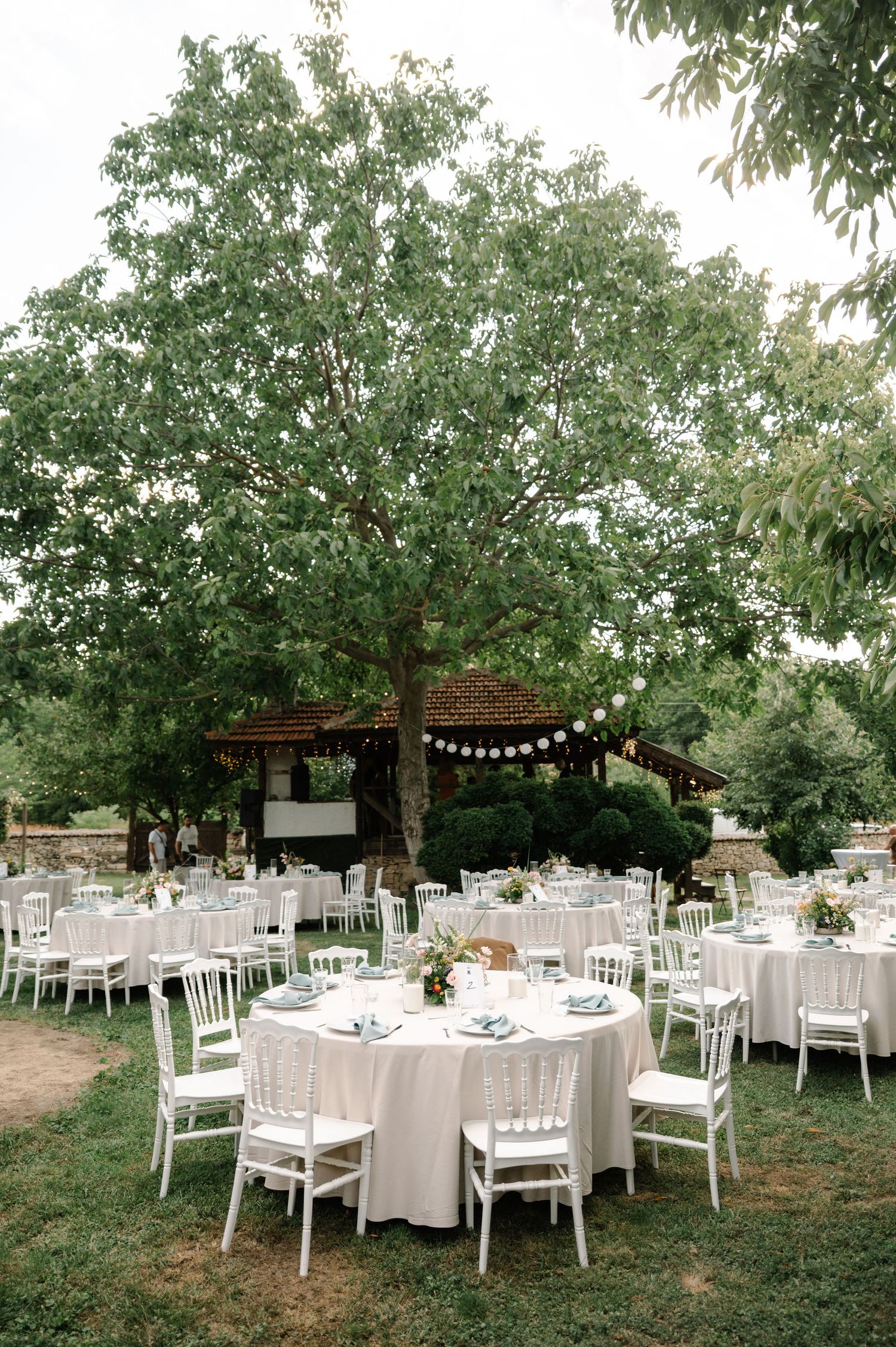 Elegant outdoor wedding reception setup with white tables and chairs under string lights and lush green trees.