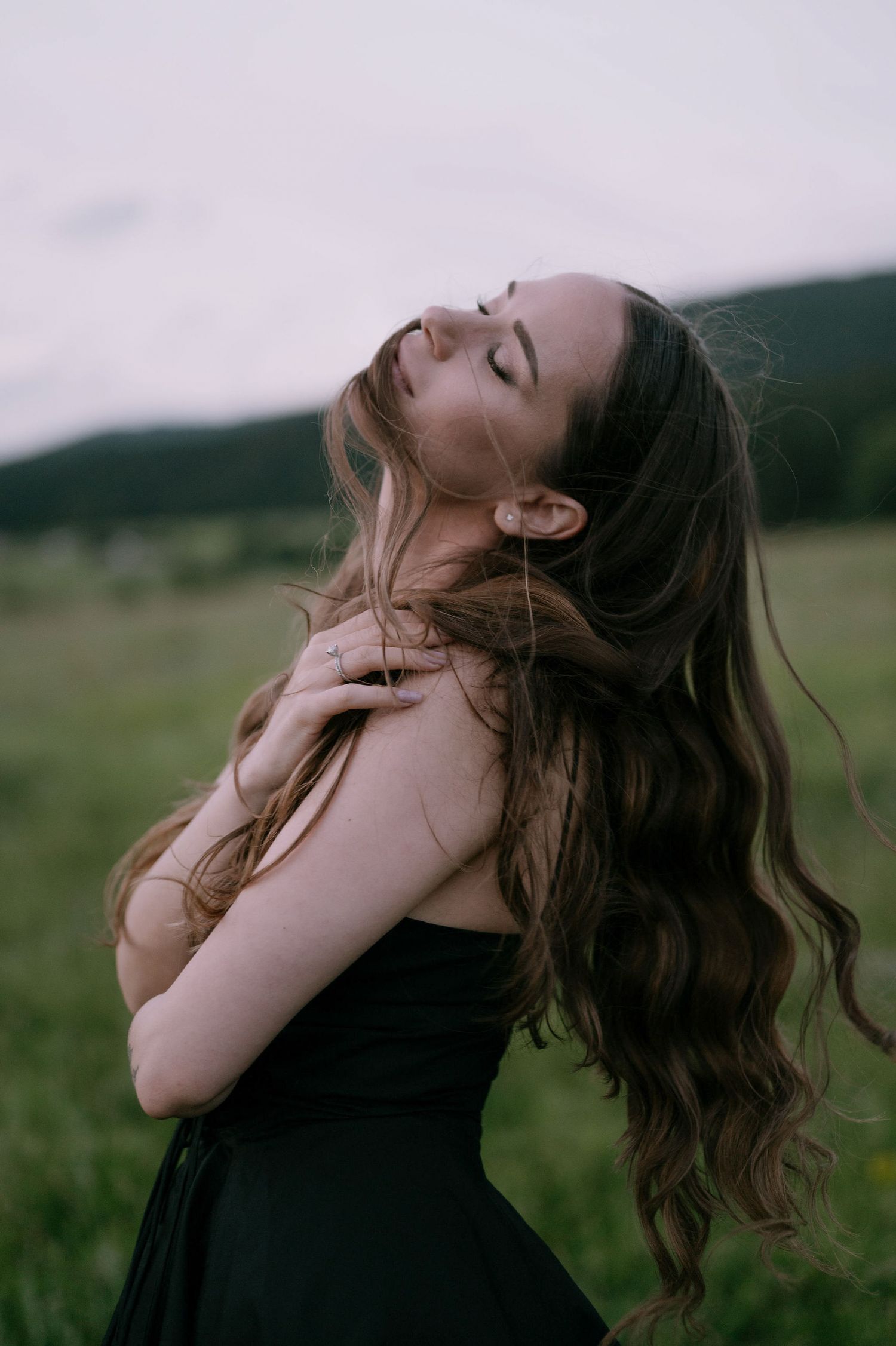 A person in a black dress tilts their head back with flowing long brown hair against a moody outdoor landscape.