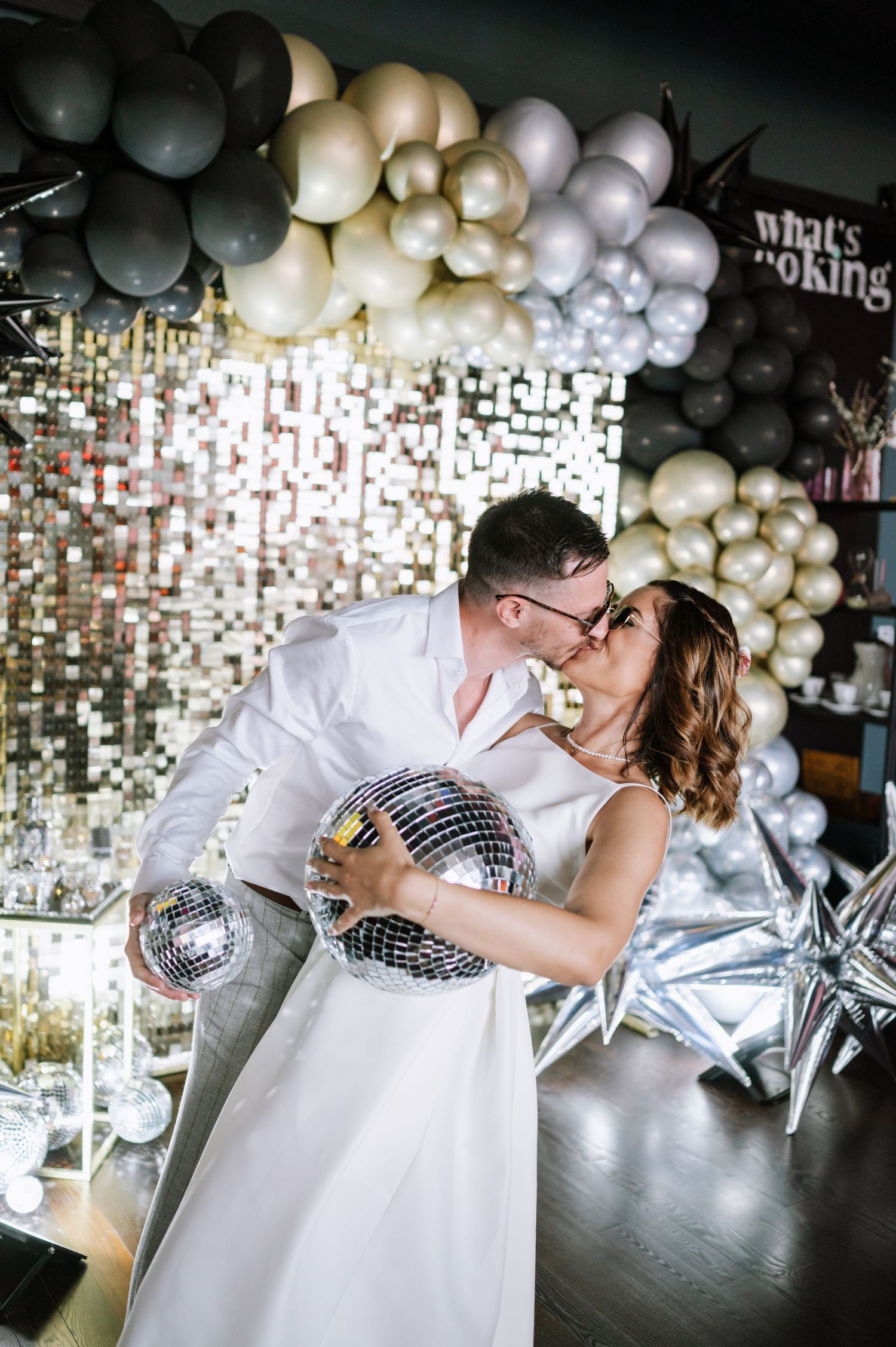 Romantic couple embraces under silver and gold balloon decorations in a stylish venue with disco ball accents.