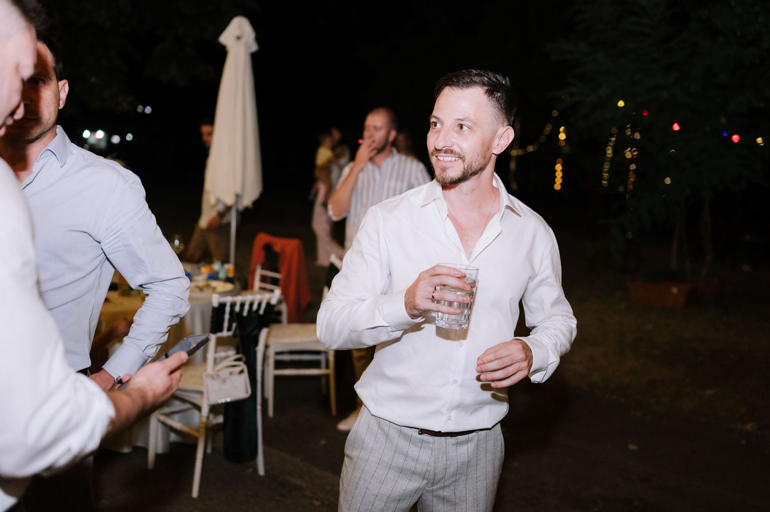 A person in a white button-down shirt laughs while holding a drink at an outdoor evening event.