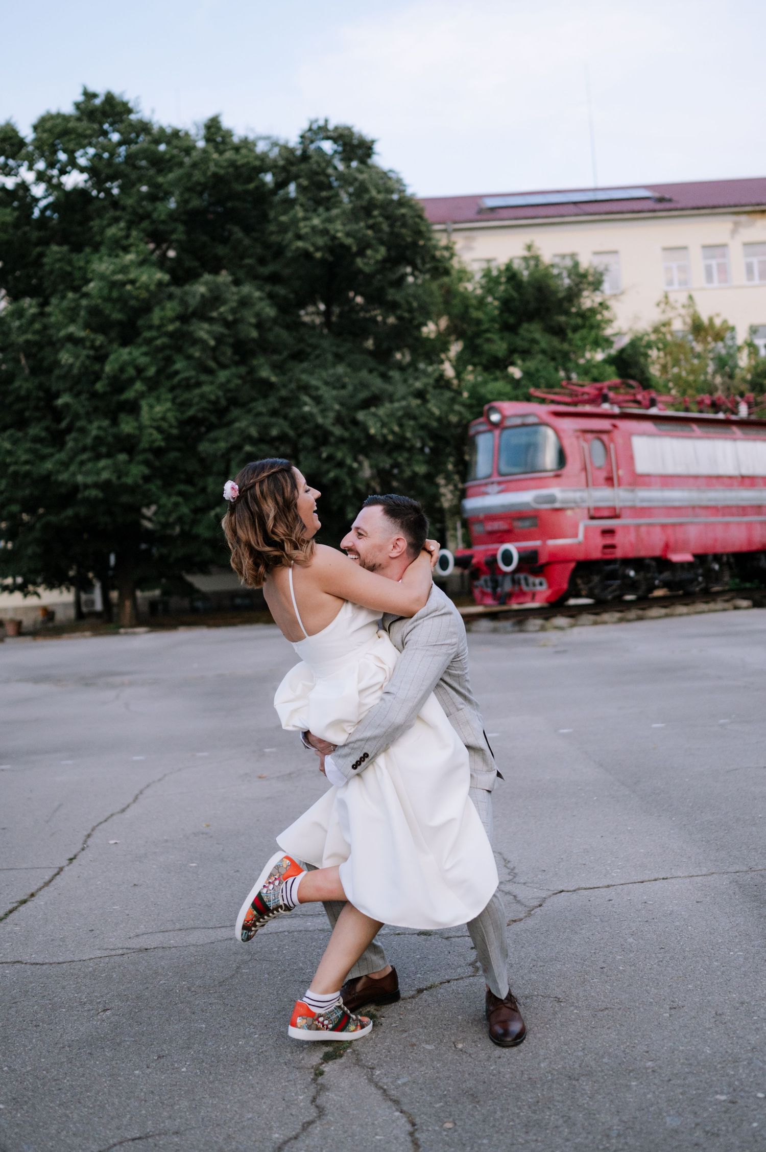A couple in wedding attire playfully dances and poses together in a parking lot with a red train car in the background.