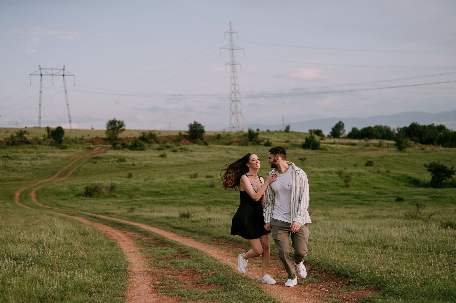 A couple walks together on a winding dirt path through a green field with power lines in the distance at sunset.