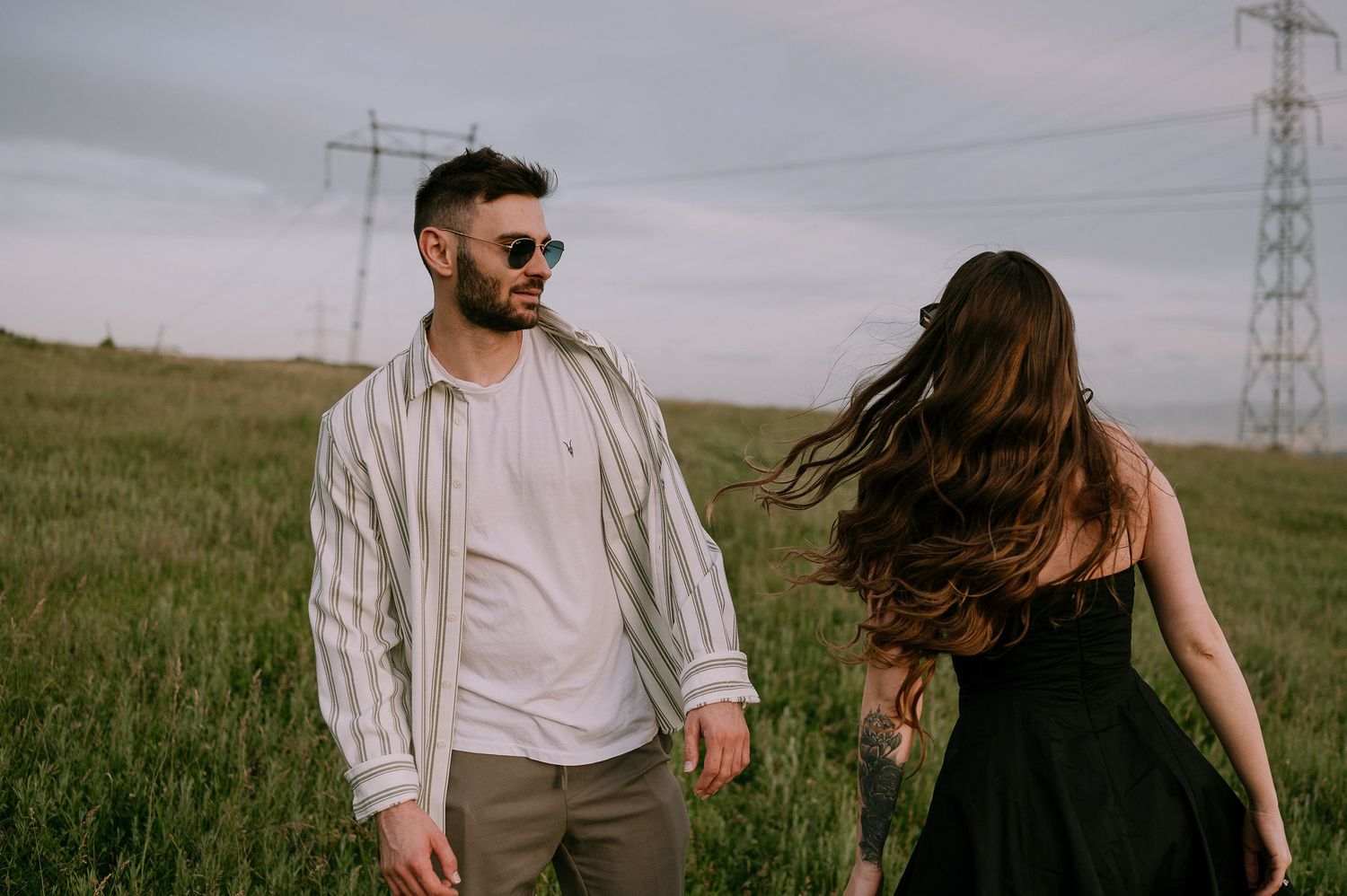 A romantic couple walks through a grassy field with power lines in the background during golden hour.