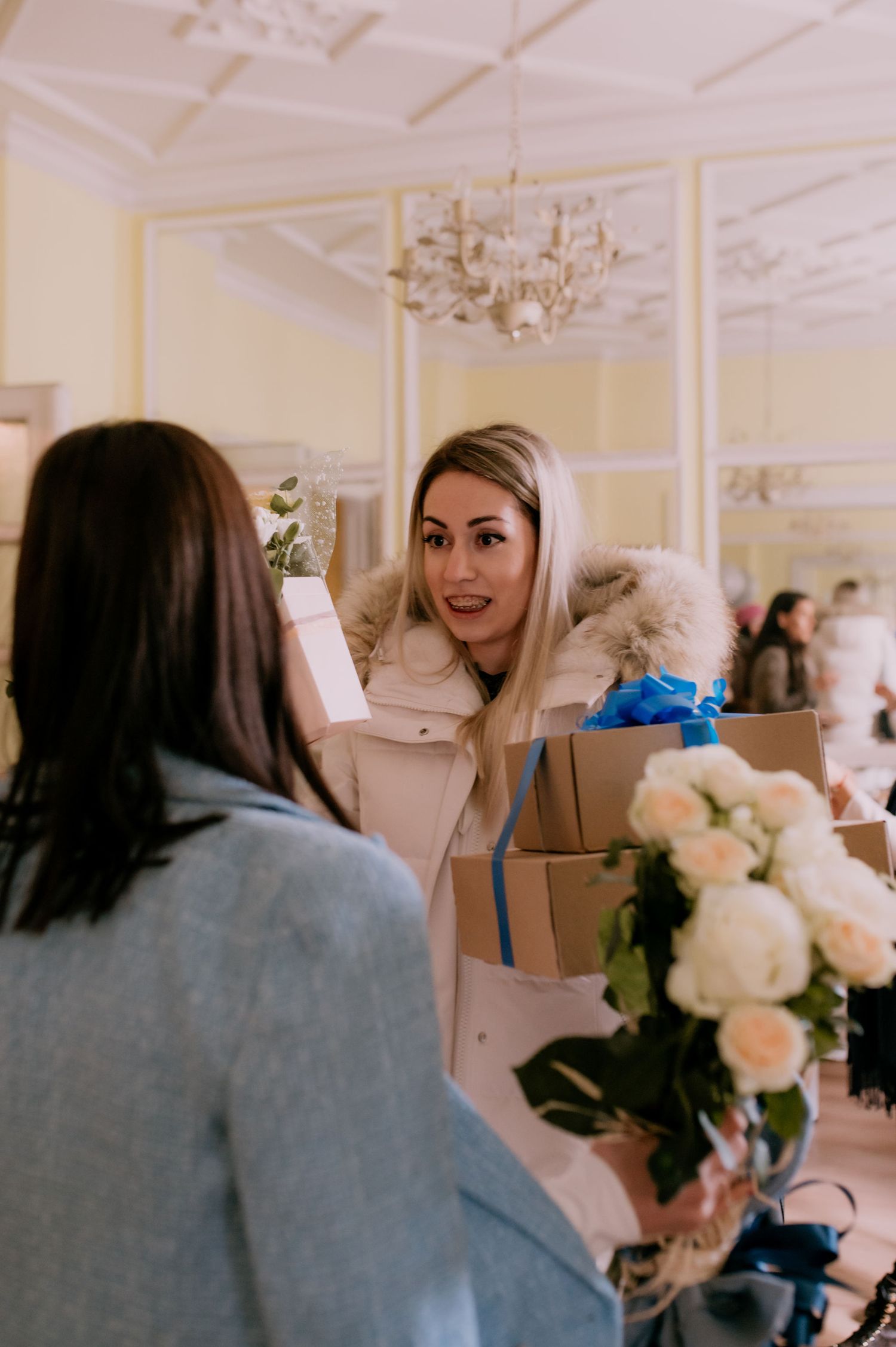 Two people engage in conversation while holding bouquets of white roses in an elegant room with ornate moldings.
