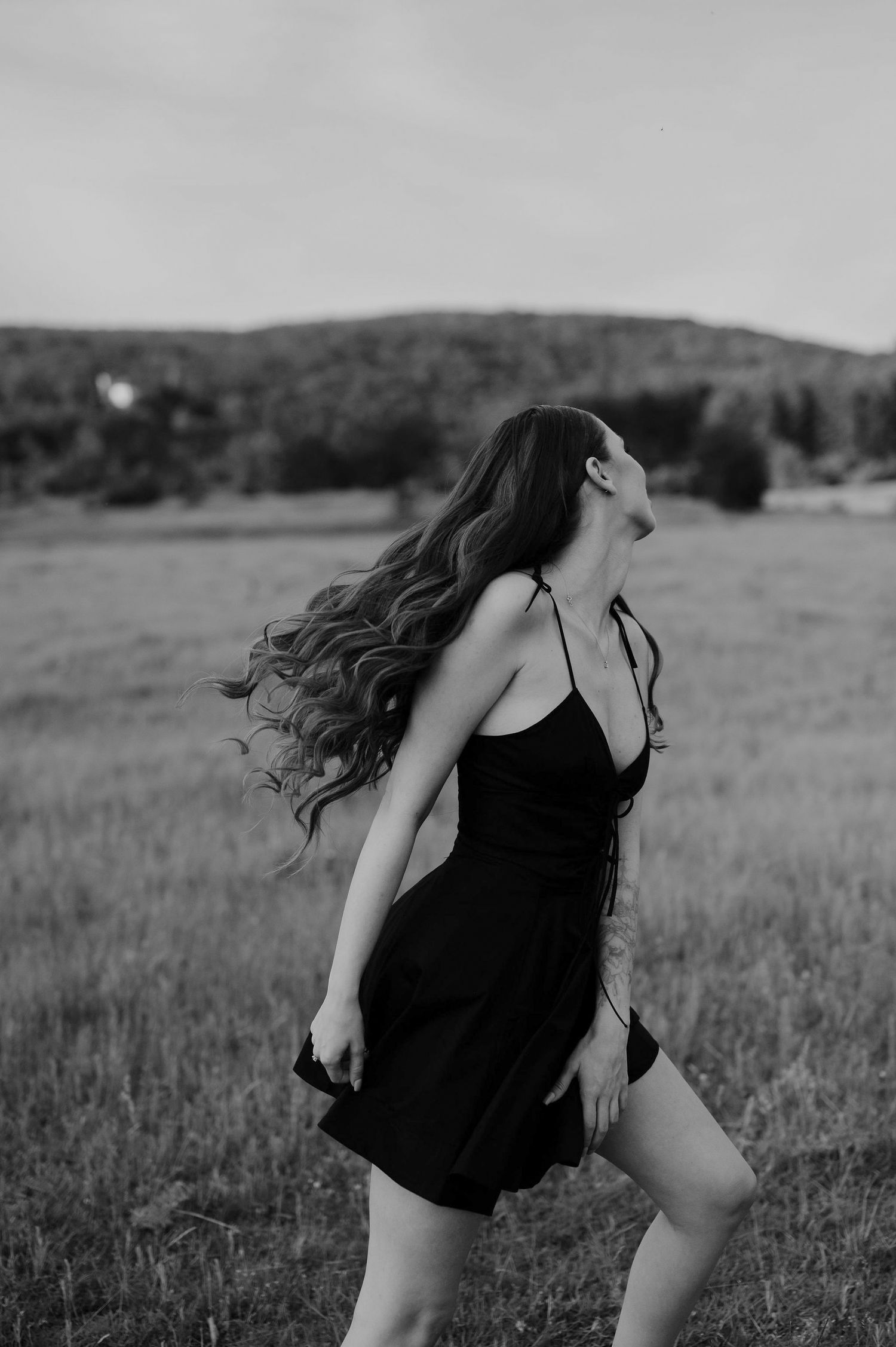 A person in a black dress dances freely in a grassy meadow with mountains in the background, captured in black and white.
