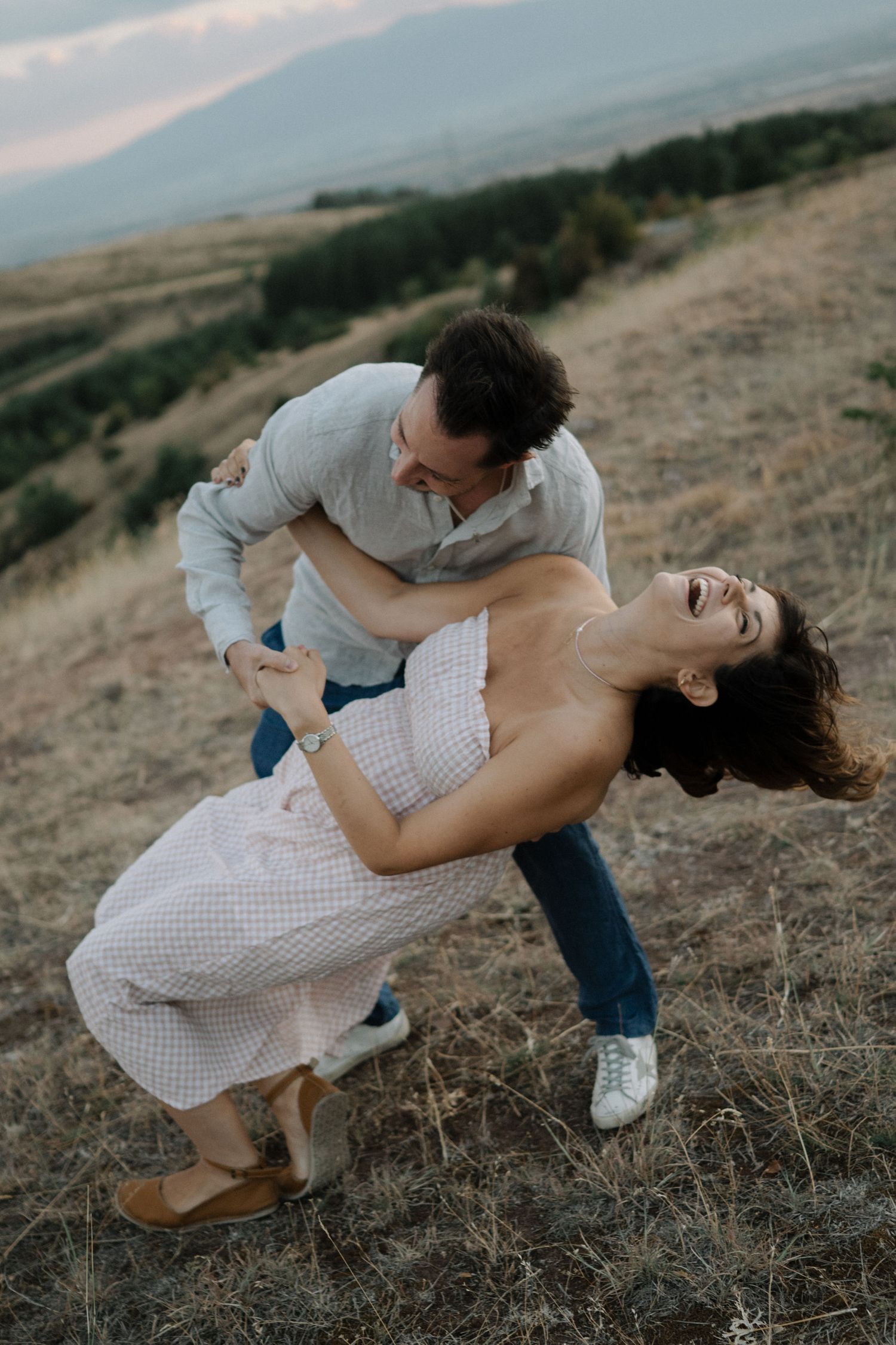 A couple in casual attire dances playfully on a grassy hillside during golden hour.
