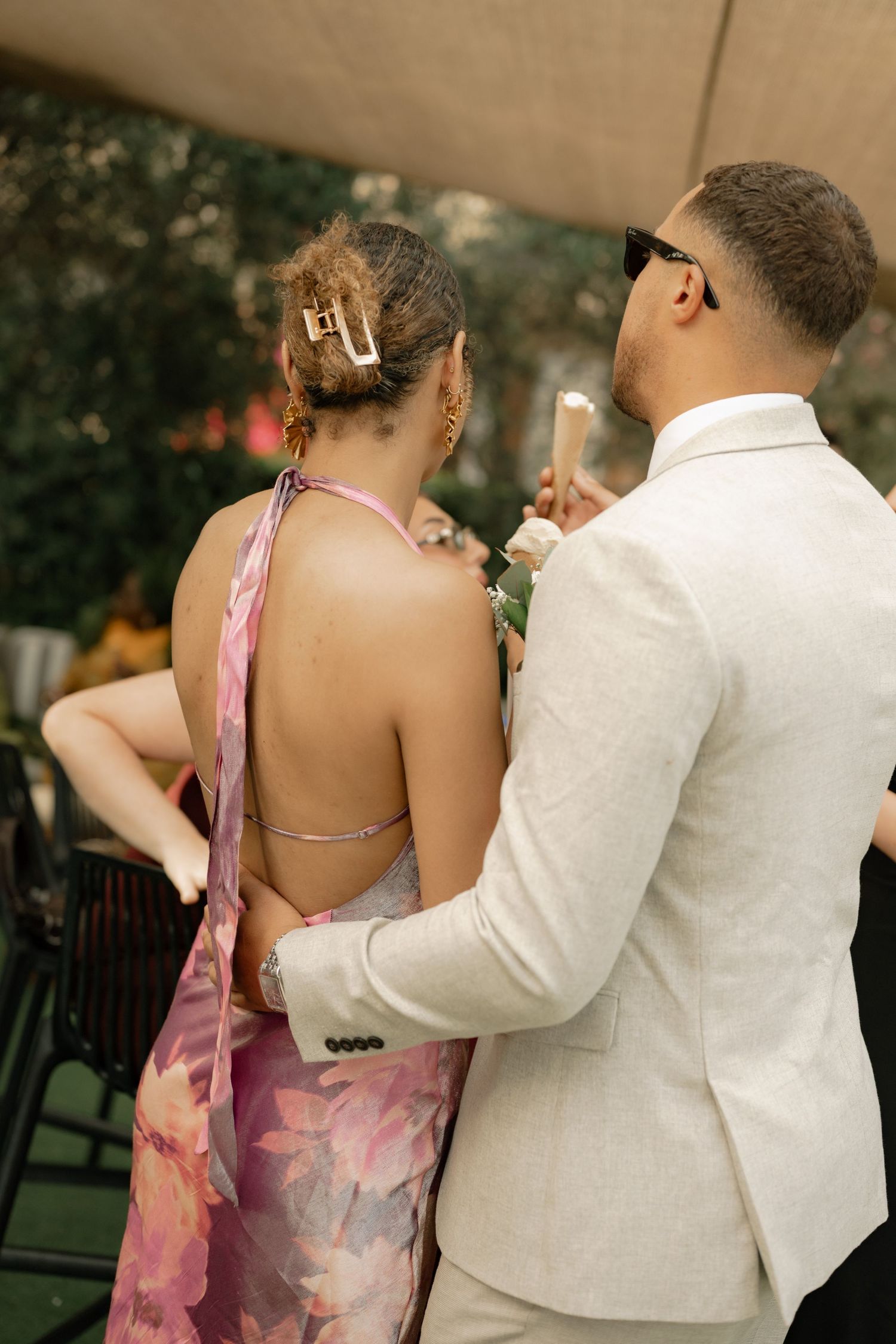 Couple at formal event, woman in backless floral halter dress, man in white suit holding champagne glass.