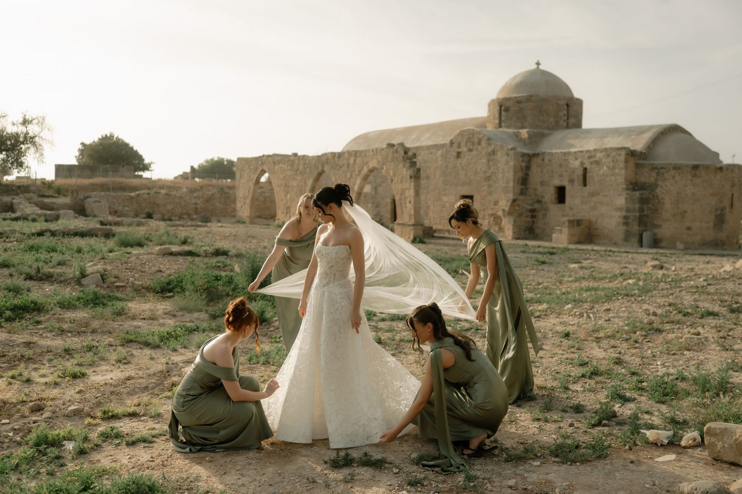 Bride in white gown with bridesmaids in sage green dresses adjusting veil near ancient stone ruins outdoors.