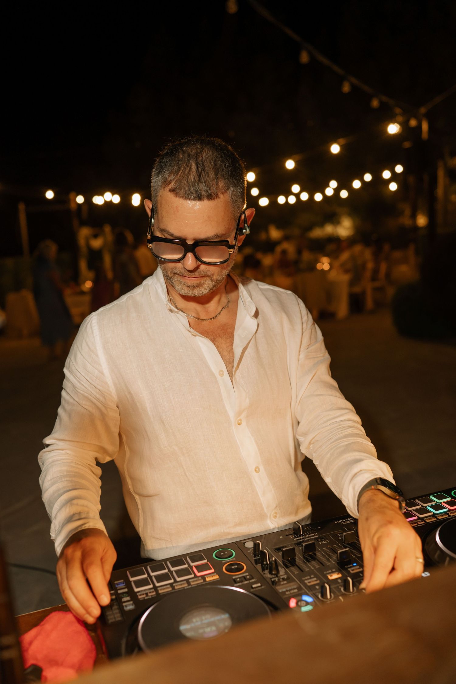 Middle-aged male DJ wearing glasses and white linen shirt mixing music at outdoor evening event with string lights.