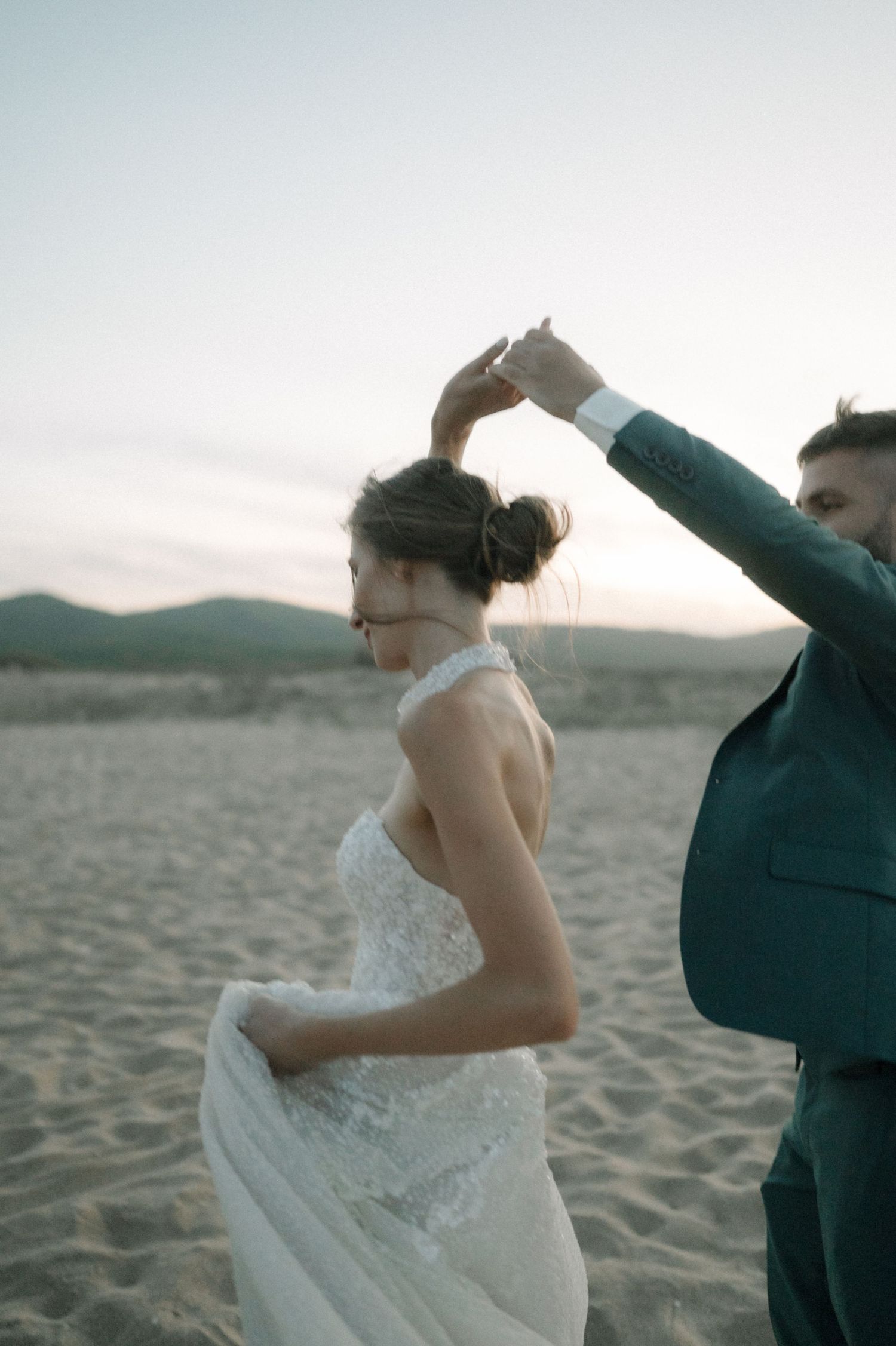 A bride in a lace gown and groom in a teal suit dance on a sandy beach at sunset.