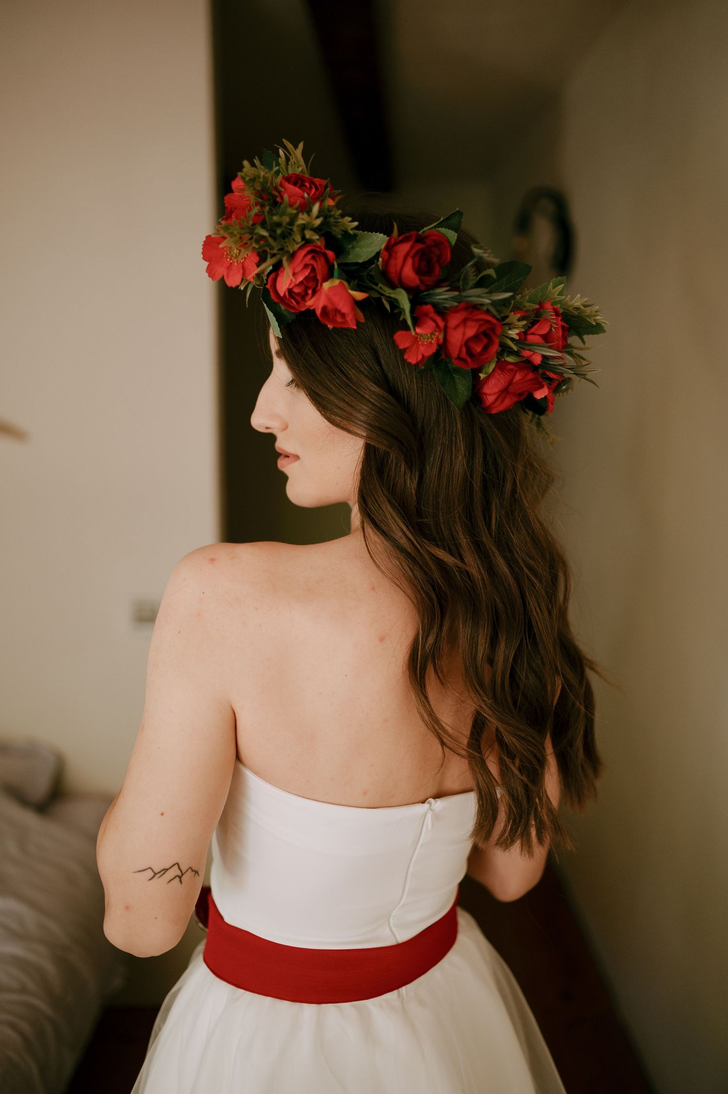 Back view of a white wedding dress with red sash and floral crown featuring red roses.