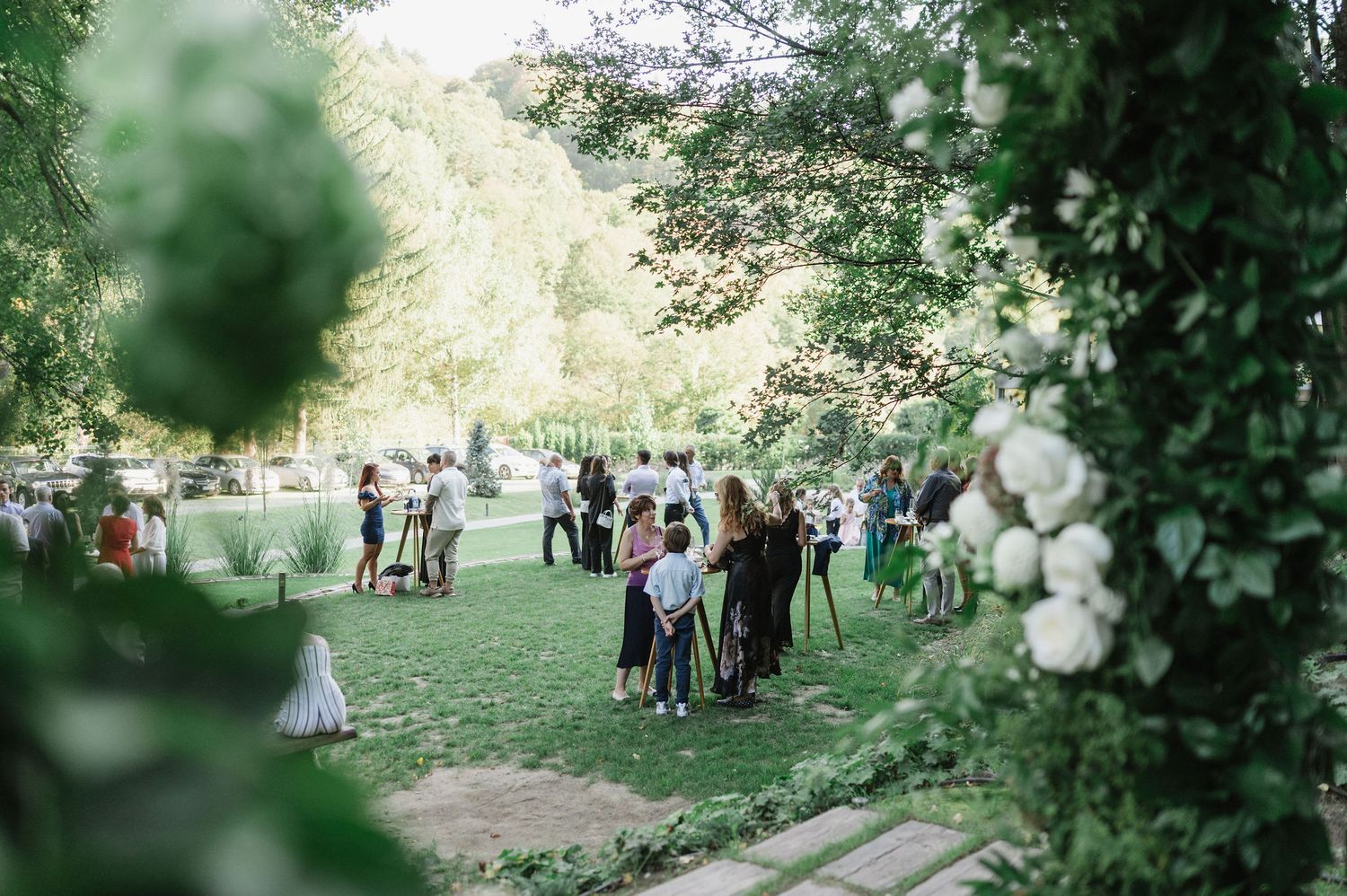 Guests gather outdoors for a celebration in a wooded area with white floral decorations.