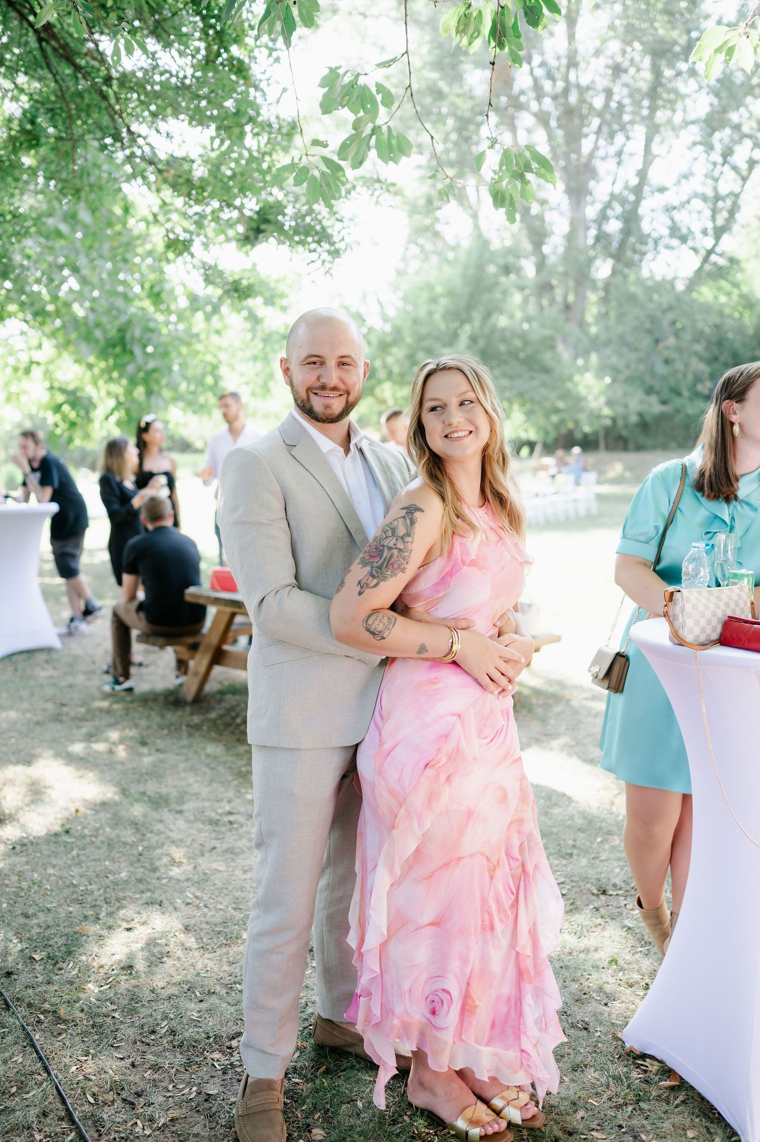 Guests enjoying a casual outdoor celebration under trees with pink and beige attire.
