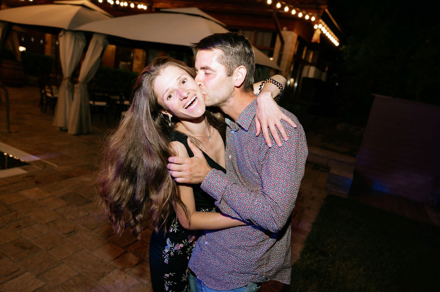 Wedding guests embrace while dancing at an outdoor evening reception under string lights.