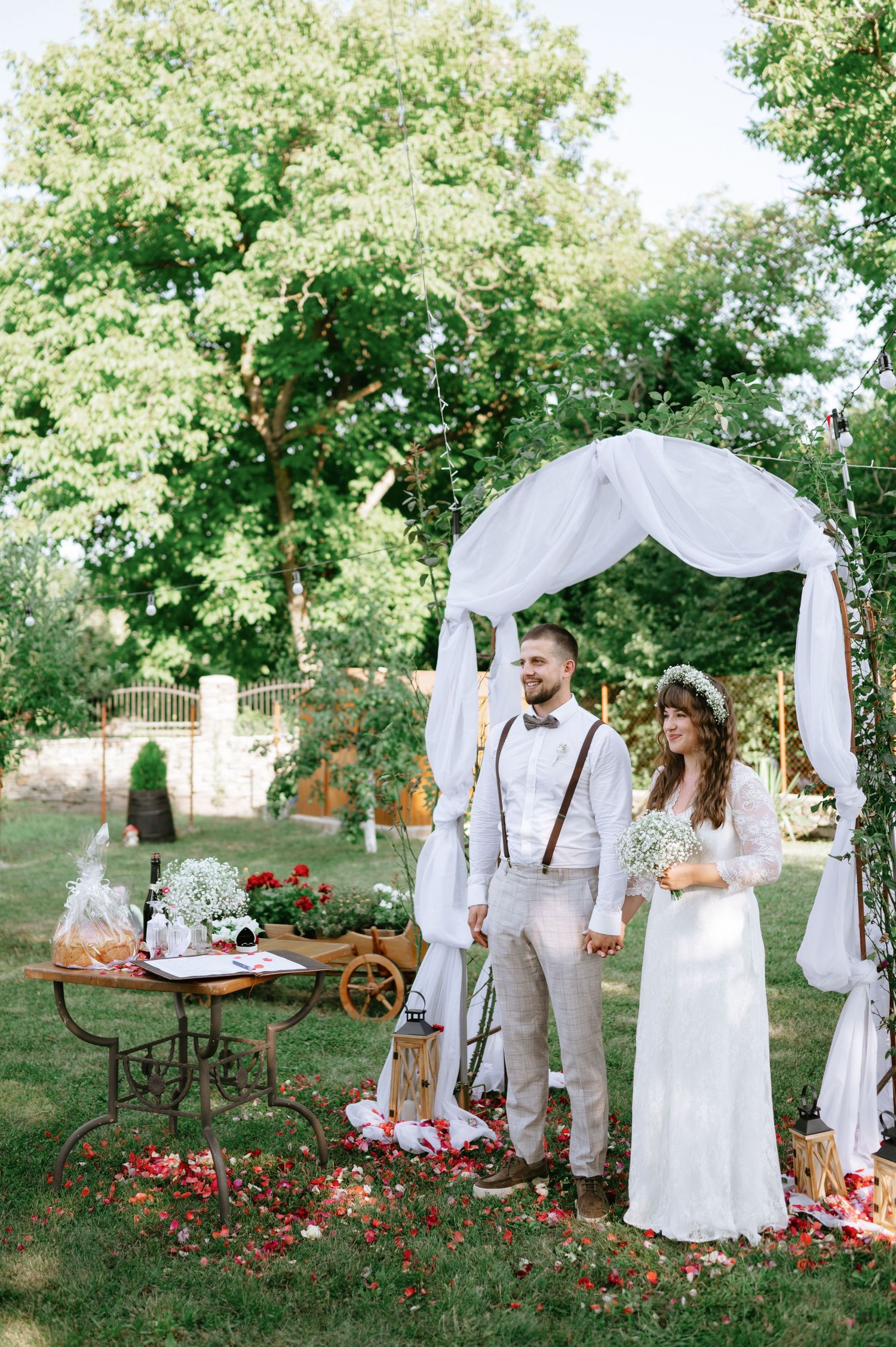 Natural garden wedding setting with white fabric arch and vintage decorations under tree canopy.