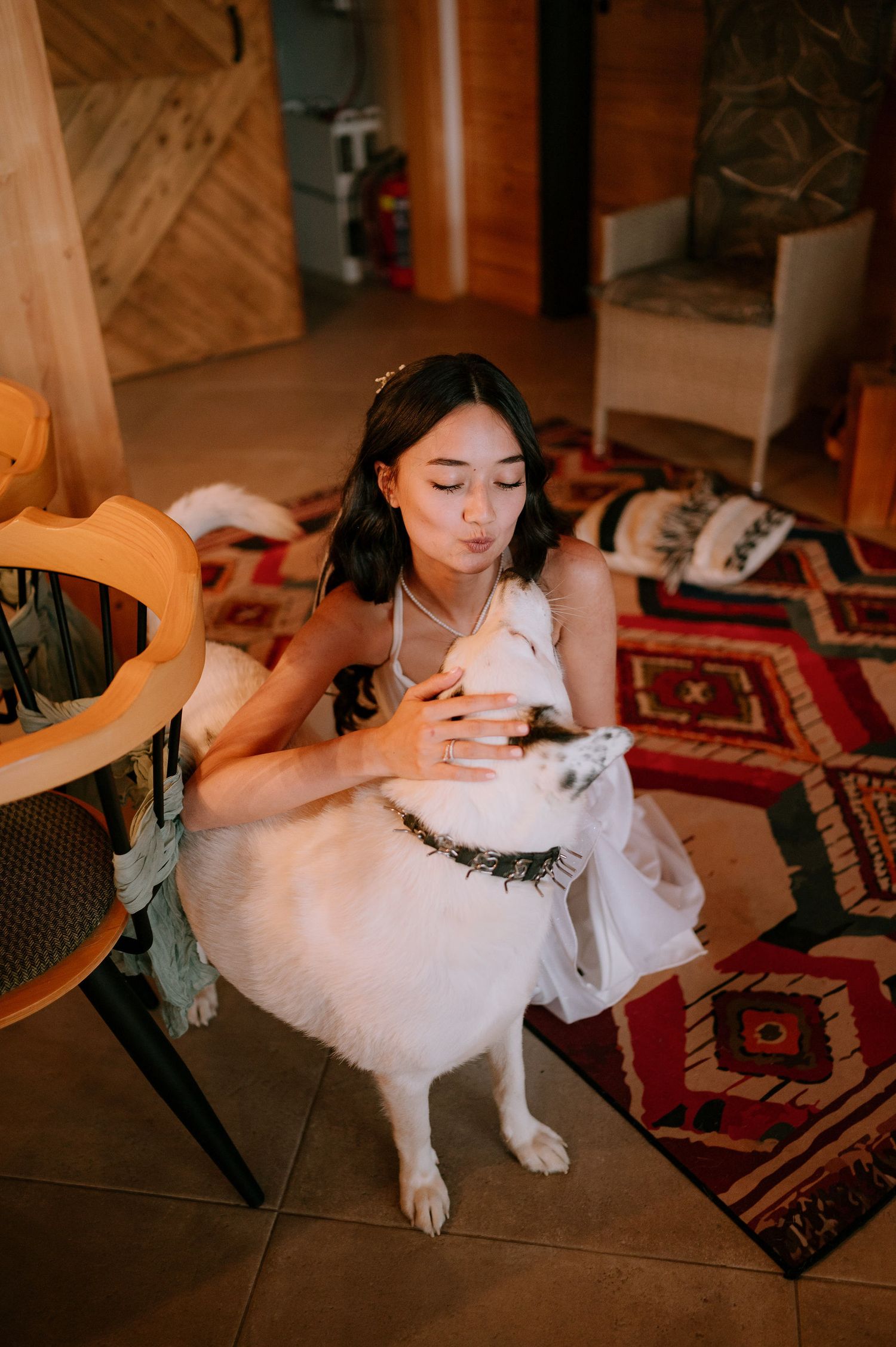 A person in white sits with a white dog on a colorful rug in an indoor setting.