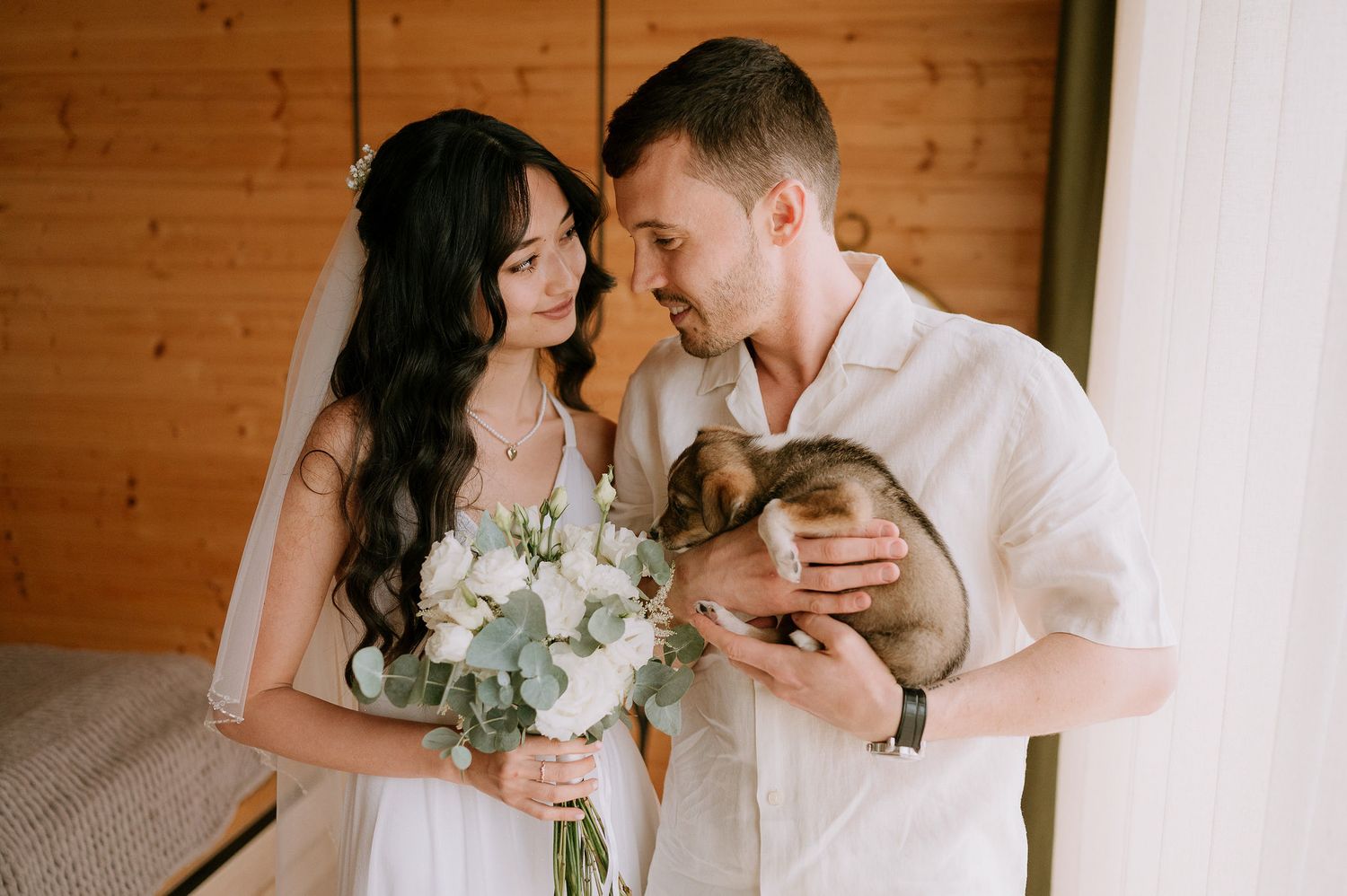 A couple in white outfits poses with their dog against a wooden wall during a romantic photoshoot.