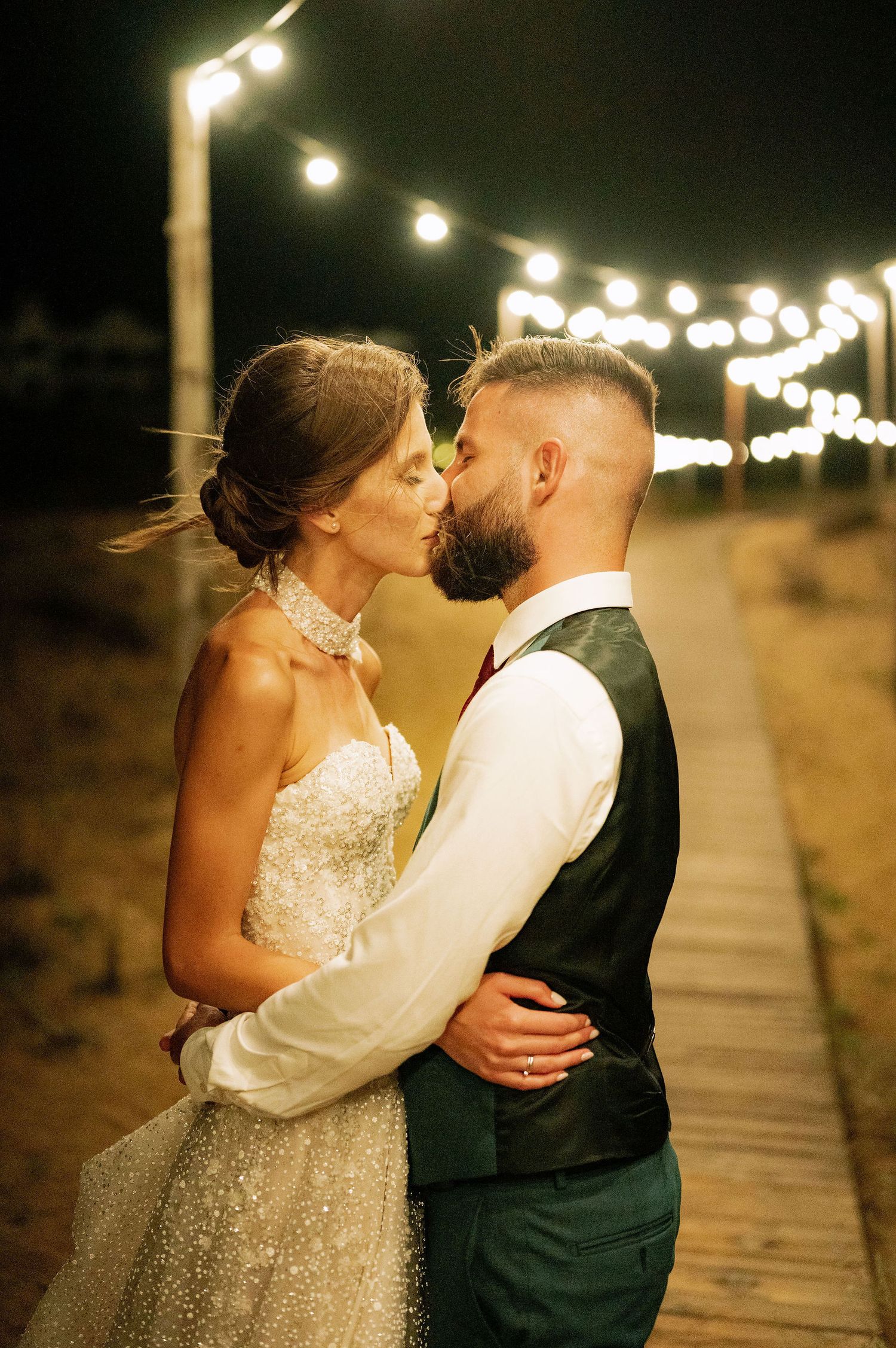 A tender moment between newlyweds beneath warm string lights on a coastal pathway.